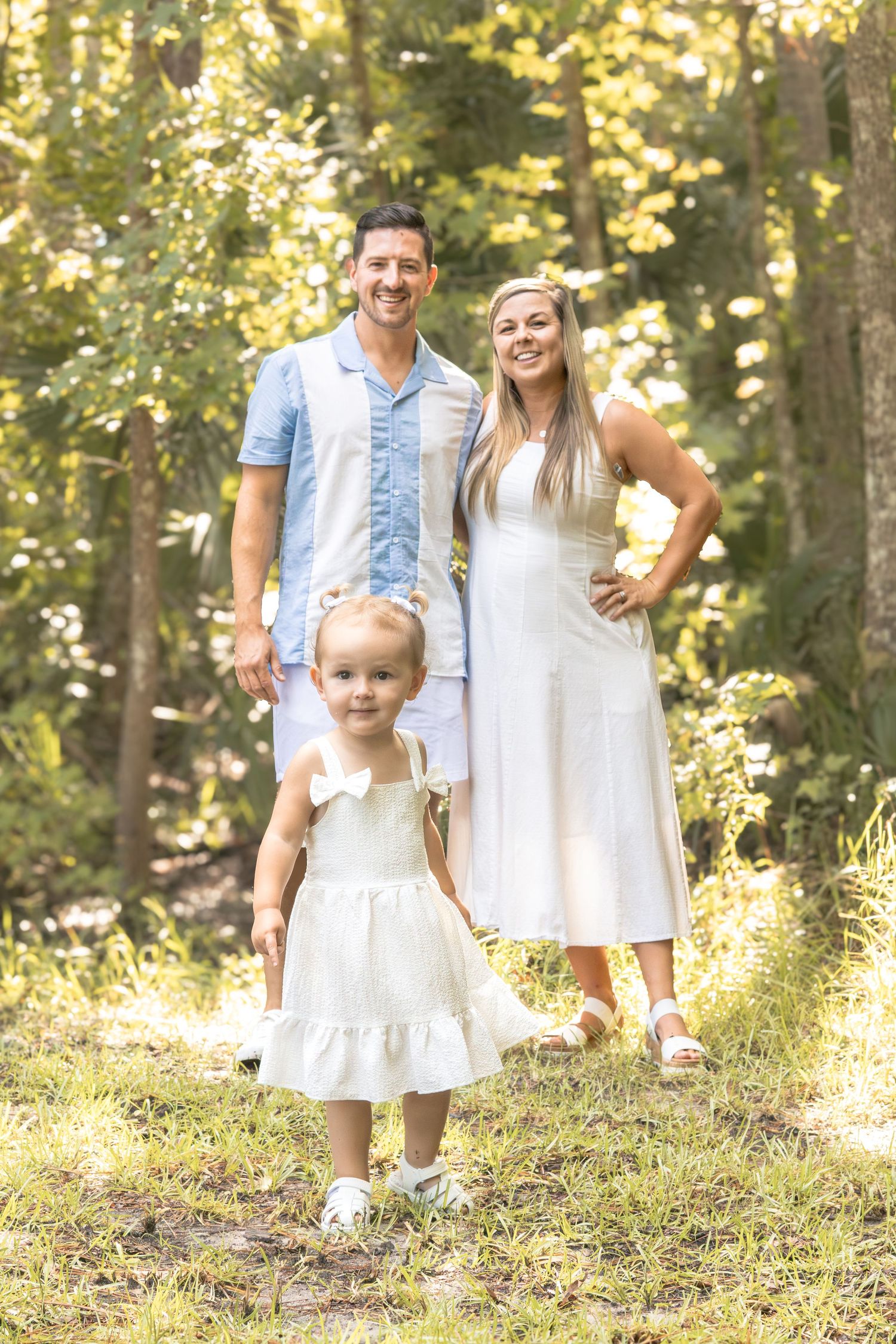 Smiling parents in light blue and white outfits stand behind their toddler daughter in a white dress in a sunny, tree-filled forest.