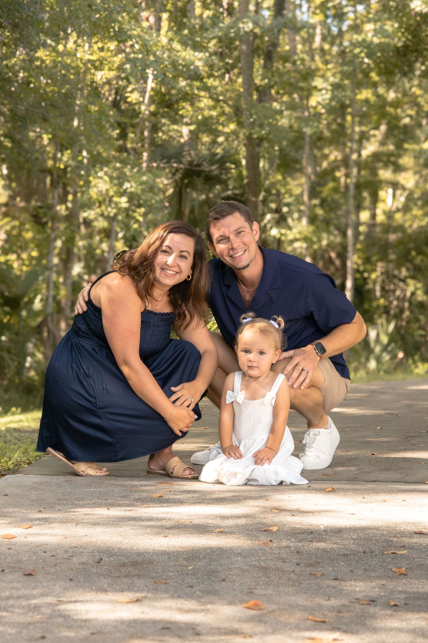 Smiling couple in navy outfits crouch on a sunny park path beside their toddler daughter in a white dress, with green trees behind them.