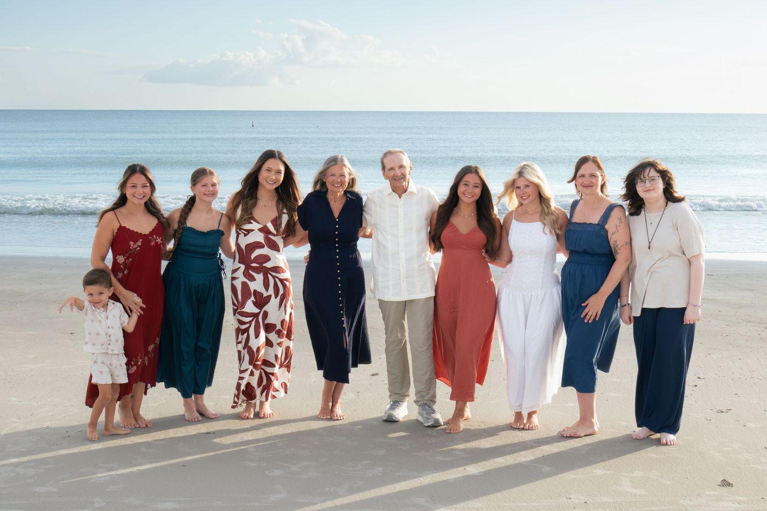 Large family in coordinated summer outfits stands together on the sand with a small child, posing for a beachfront portrait.
