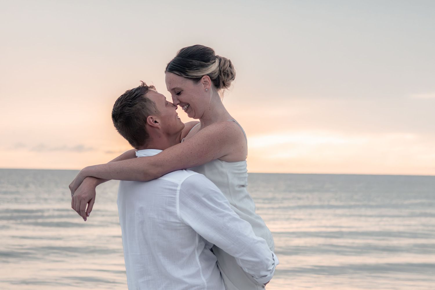 Man lifts his partner in a loving embrace at the beach as they almost kiss against a soft pastel sunset sky.