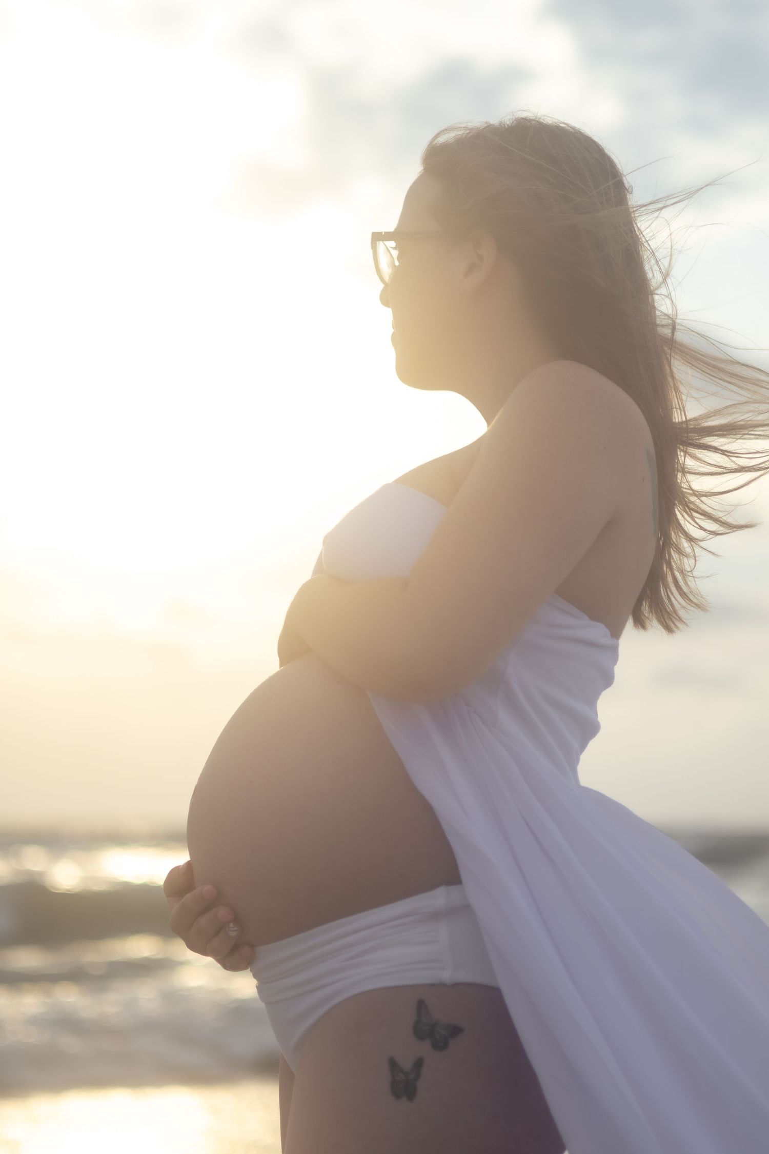 A pregnant woman with glasses and butterfly tattoos stands on the beach at sunset, her hair blowing in the breeze.