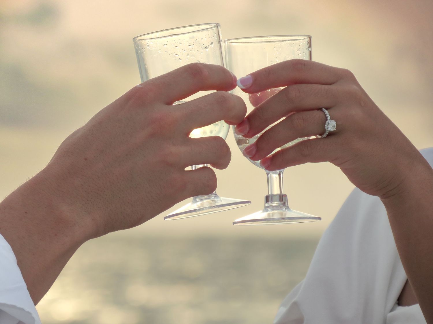 Two people clink plastic champagne glasses in a toast at sunset on the beach, one hand wearing a sparkling diamond ring.