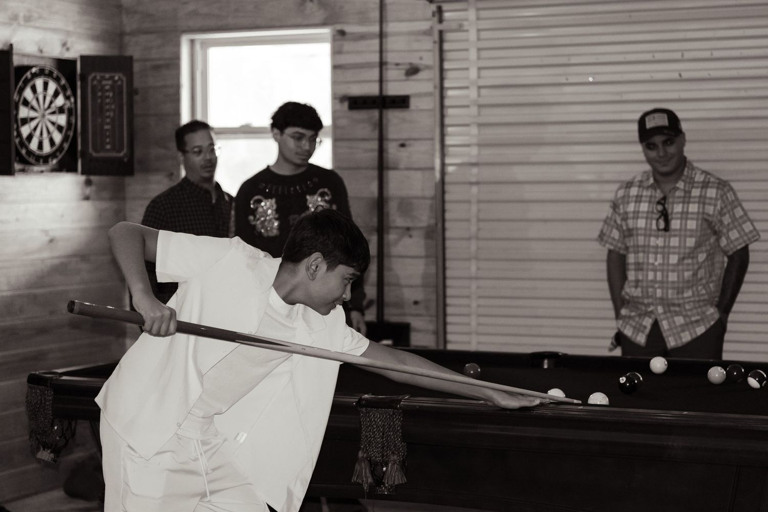 Man leans over a pool table lining up a shot while three onlookers watch near a dartboard in a game room.