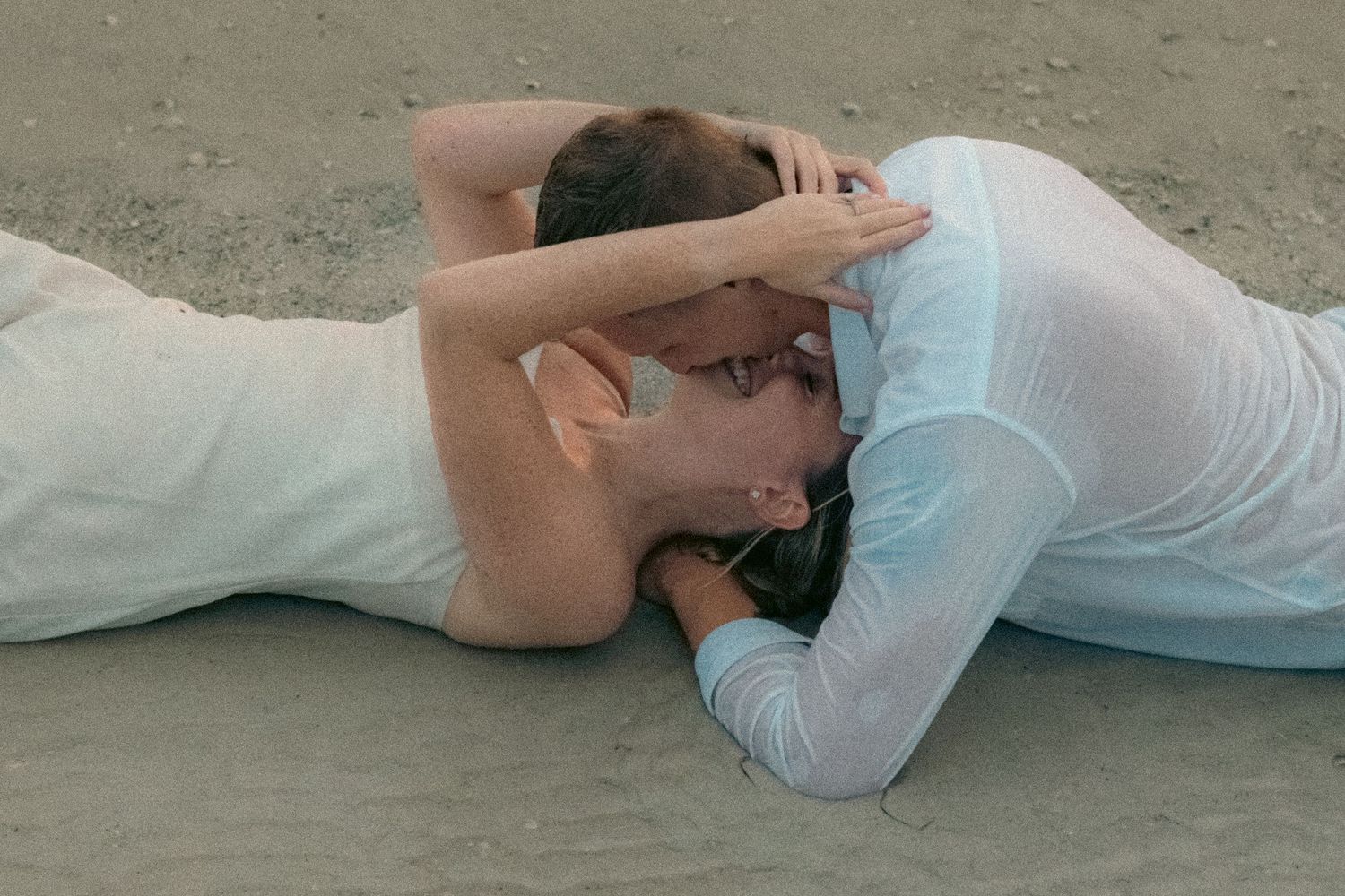 Couple in white clothing lying side by side on a sandy beach, embracing and kissing in a romantic moment.