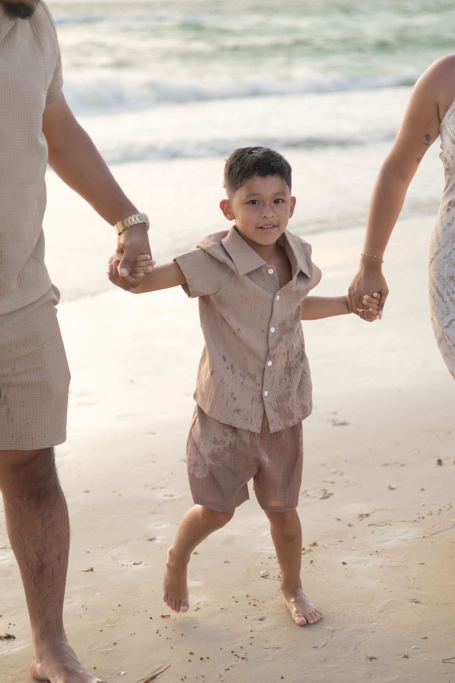 Happy young boy in a tan outfit walks barefoot on the sand while holding his parents' hands during a beach photo session.