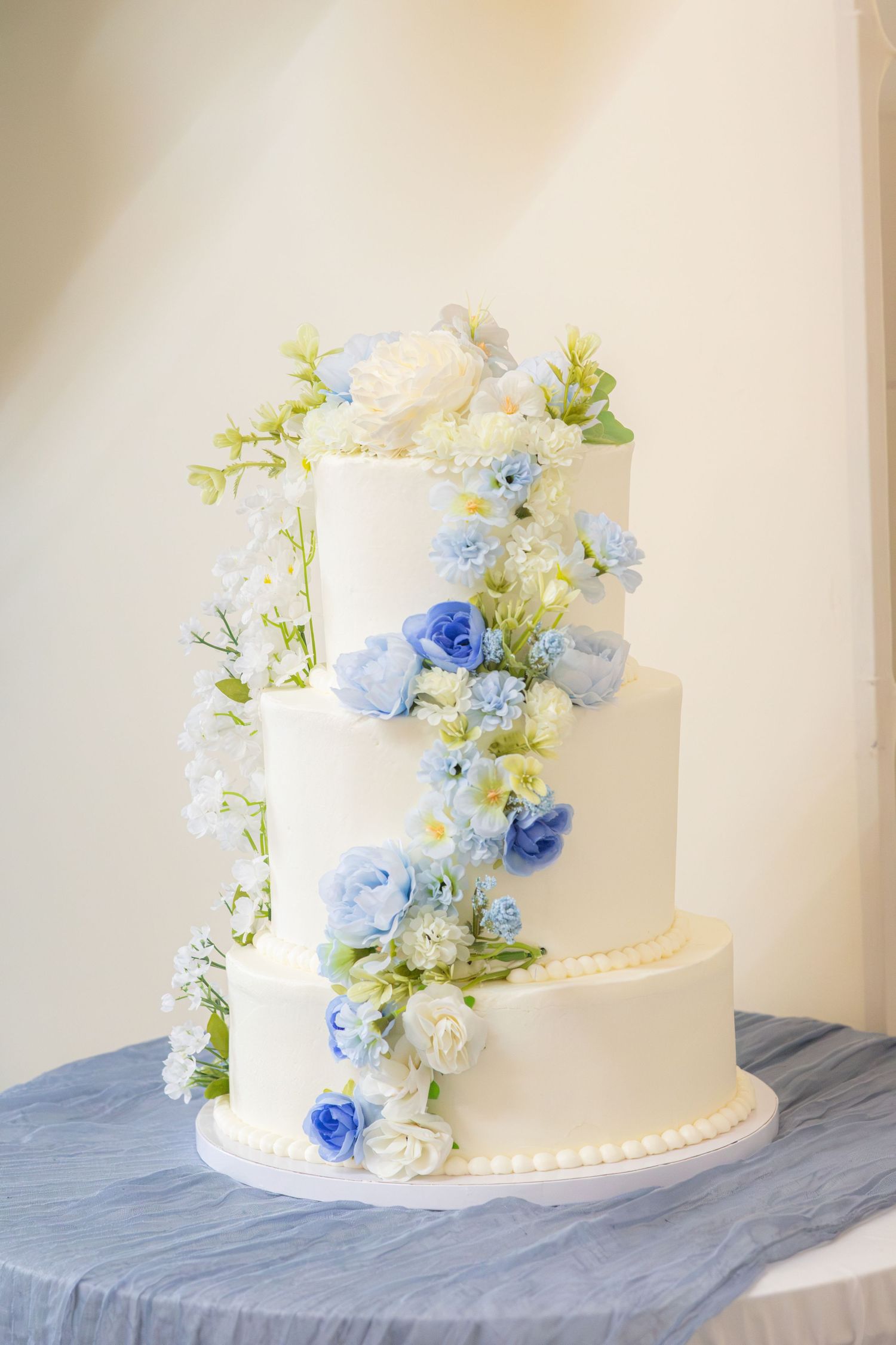 Three-tier white wedding-style cake decorated with cascading blue and white flowers on a blue draped table.