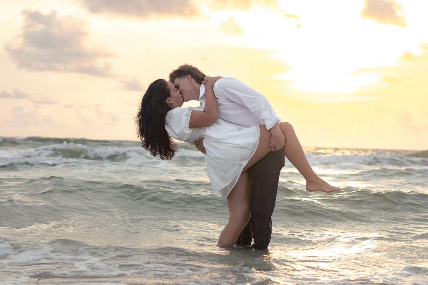 Man dipping woman in a romantic kiss in the ocean surf with a golden sunset glowing behind them.