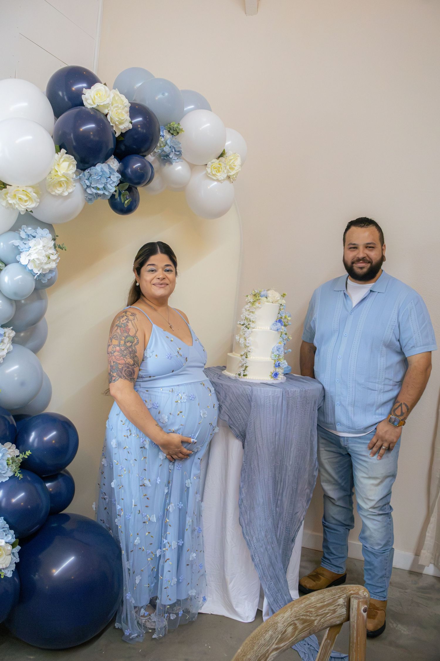 Expectant mother in a blue floral dress and her partner pose beside a white floral cake and blue balloon garland at a baby shower.
