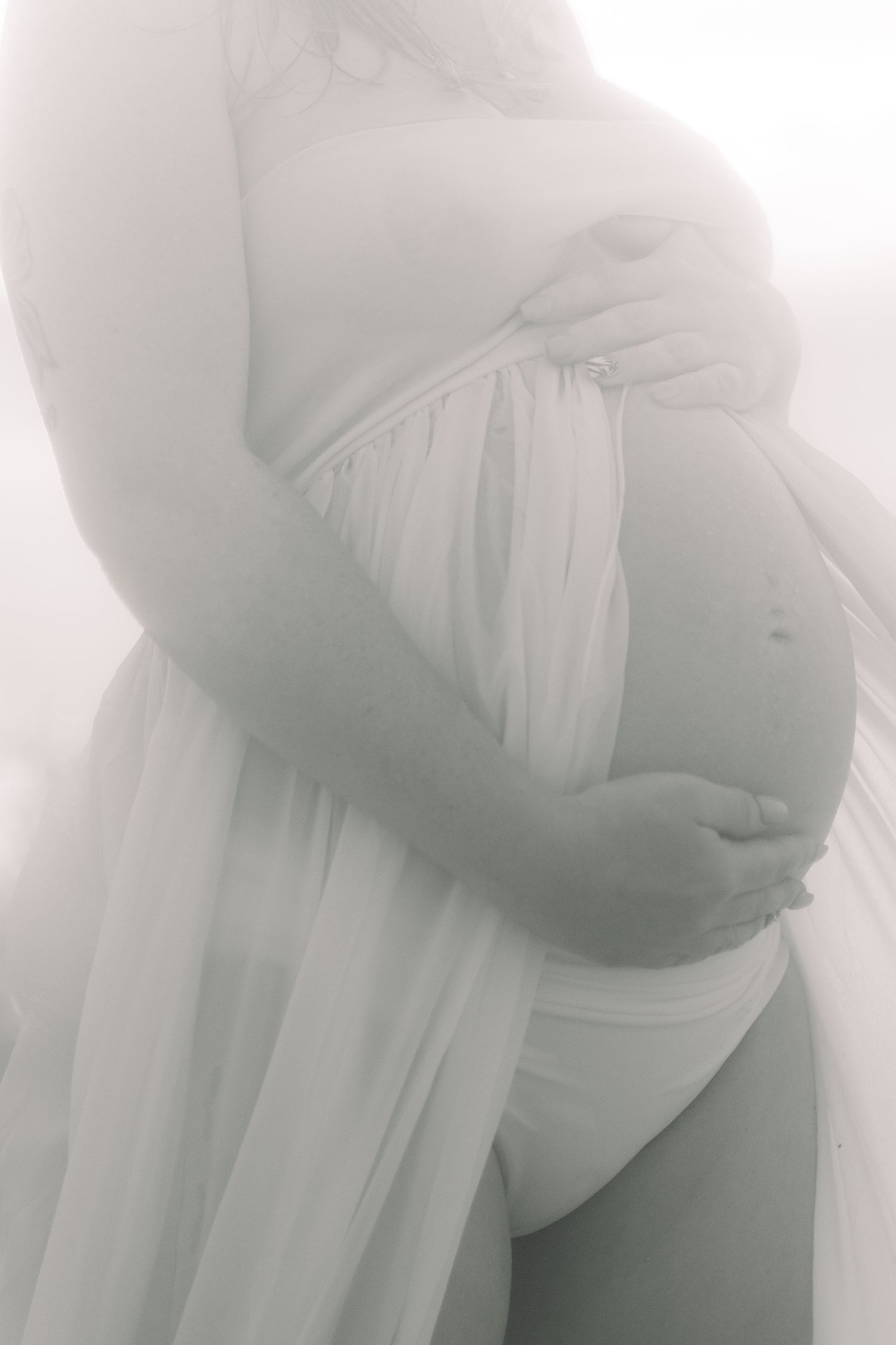 Soft, high-key black and white maternity image of a woman in sheer fabric cradling her pregnant belly.
