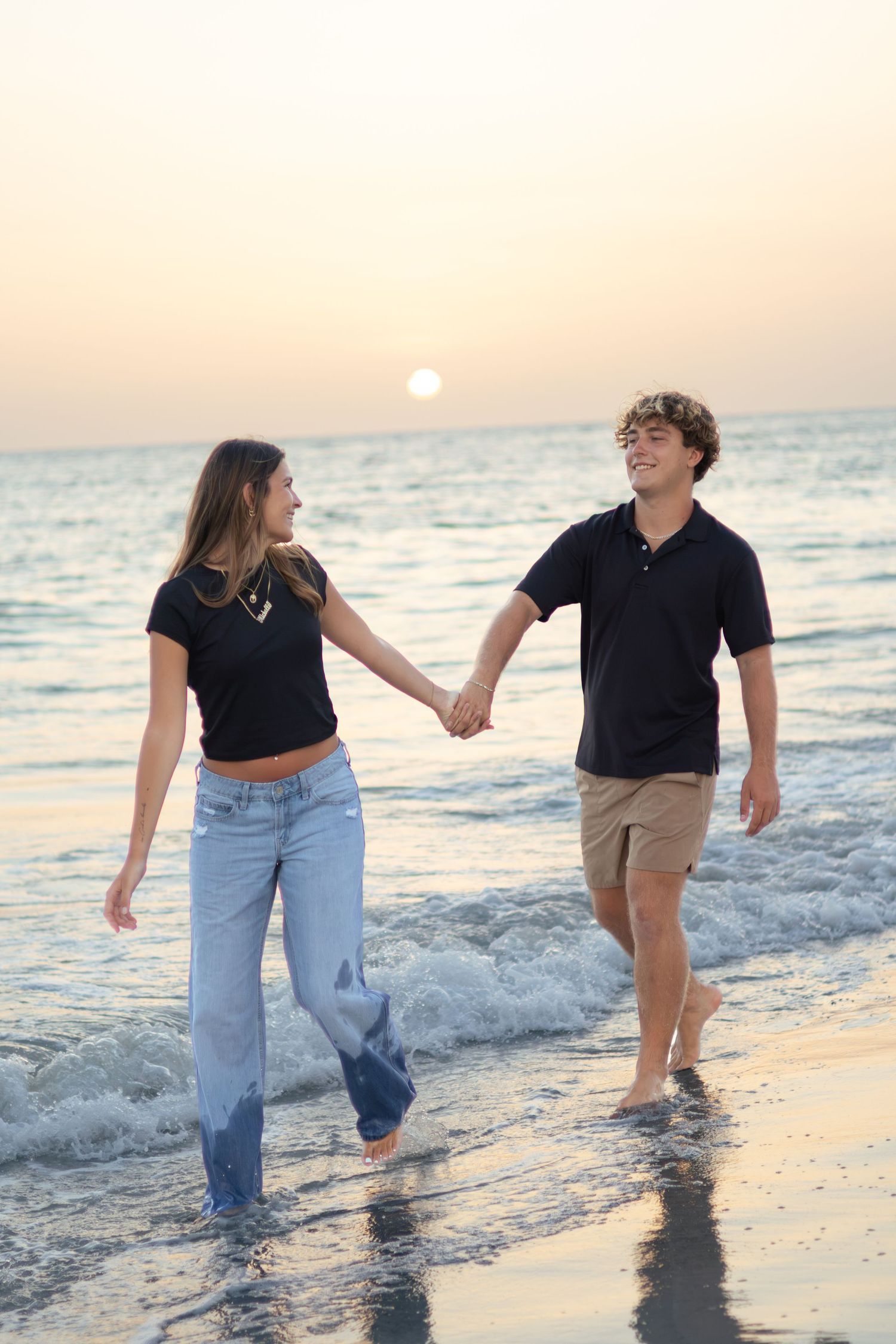 Smiling couple holds hands and walks through shallow ocean waves as the sun sets over the horizon.