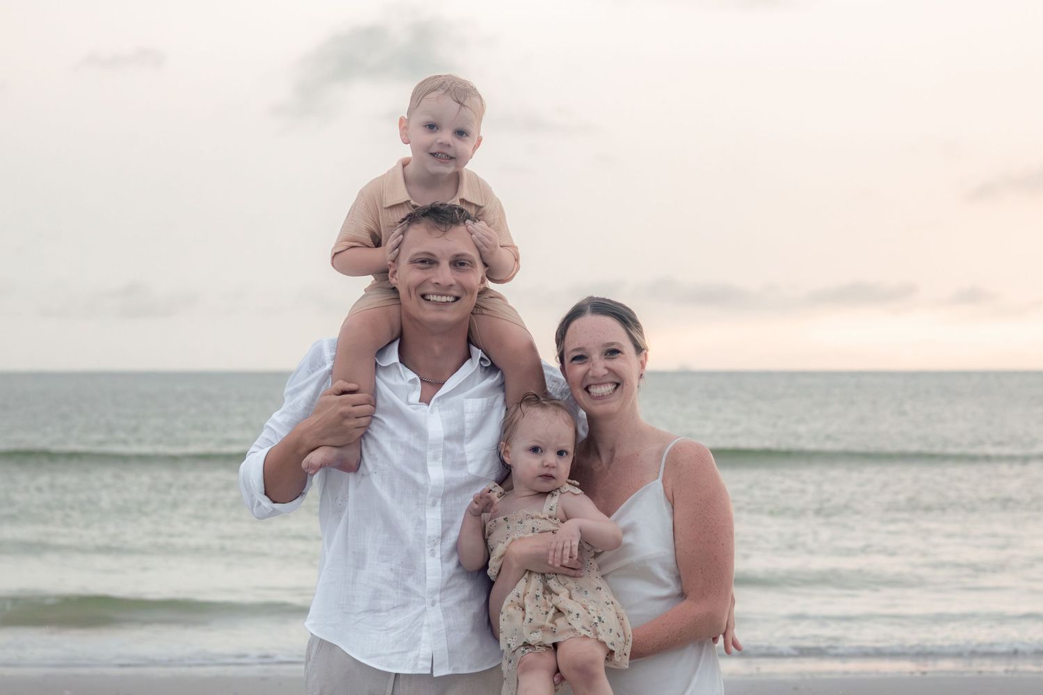 Smiling father carries a toddler on his shoulders while a mother holds a baby girl, posing together on the beach.