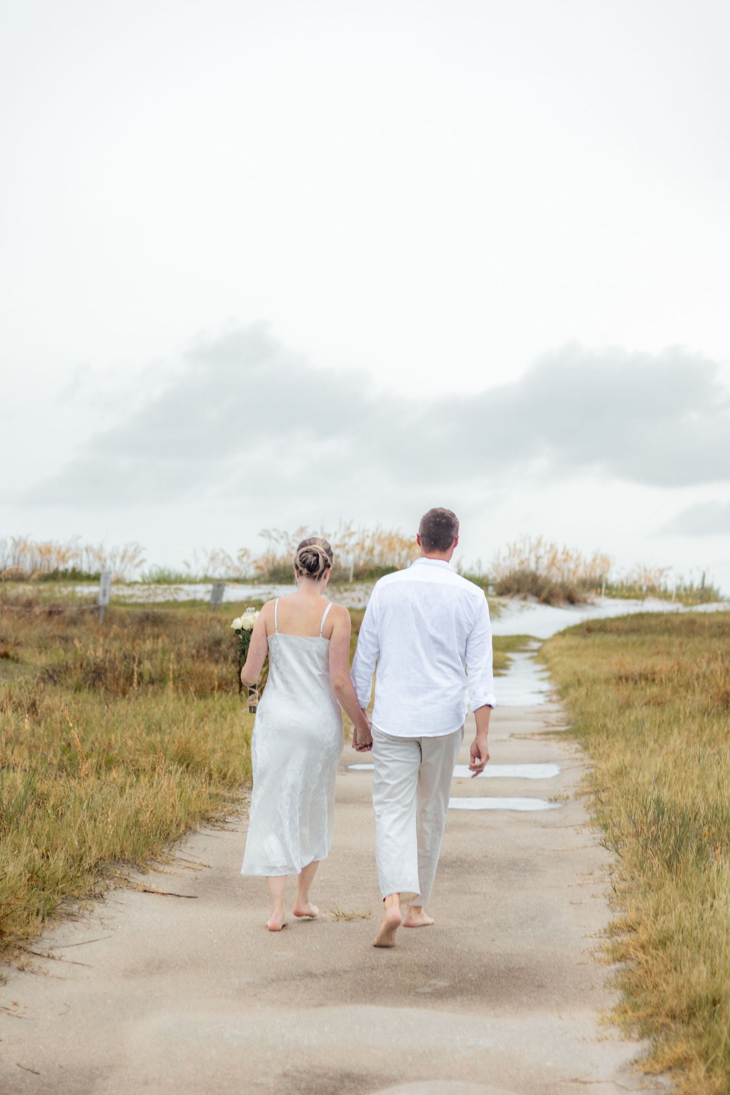 Barefoot couple in white clothing walks hand in hand along a sandy path through beach grasses.