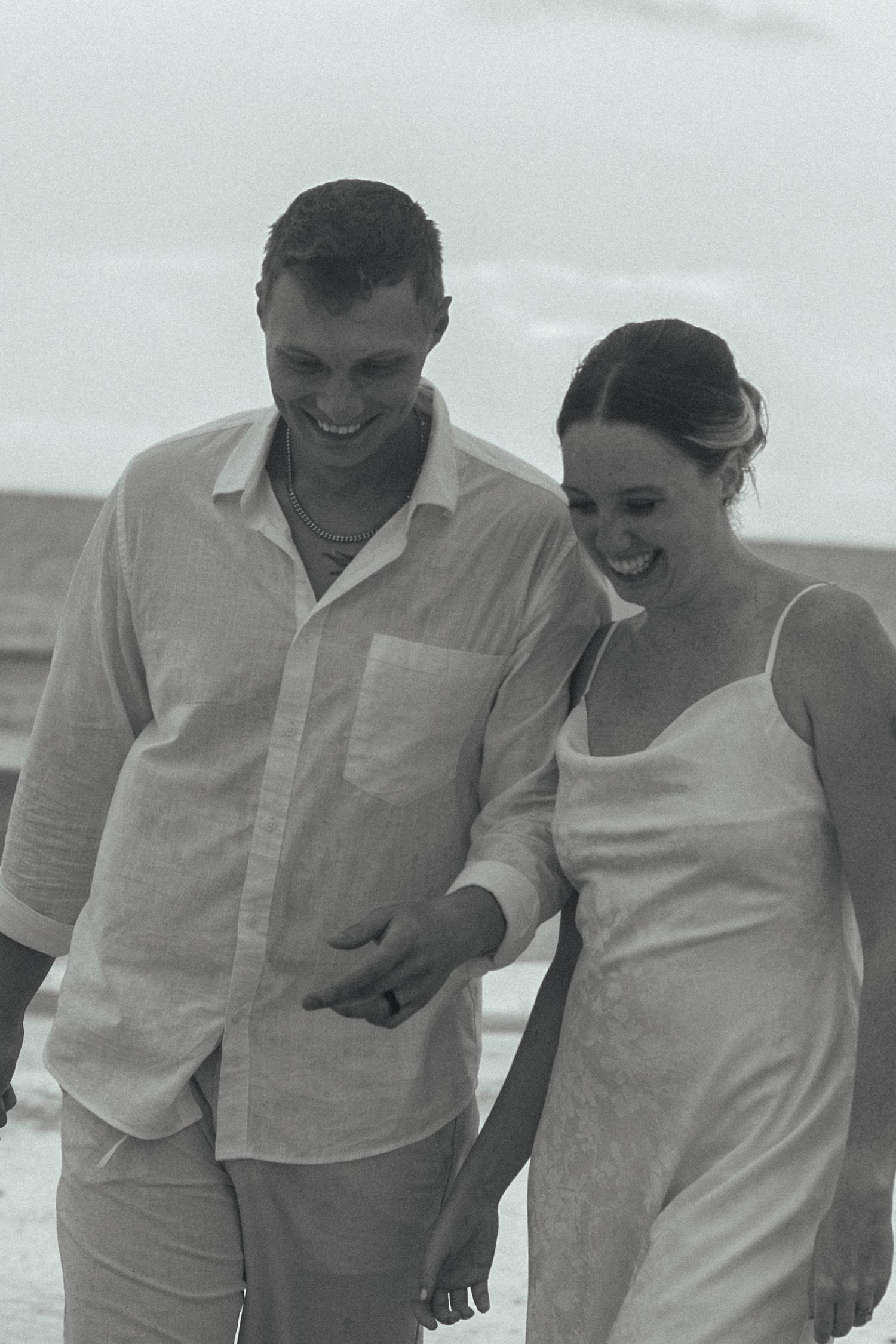 Black and white photo of a smiling couple in light linen clothing walking arm in arm along the beach.