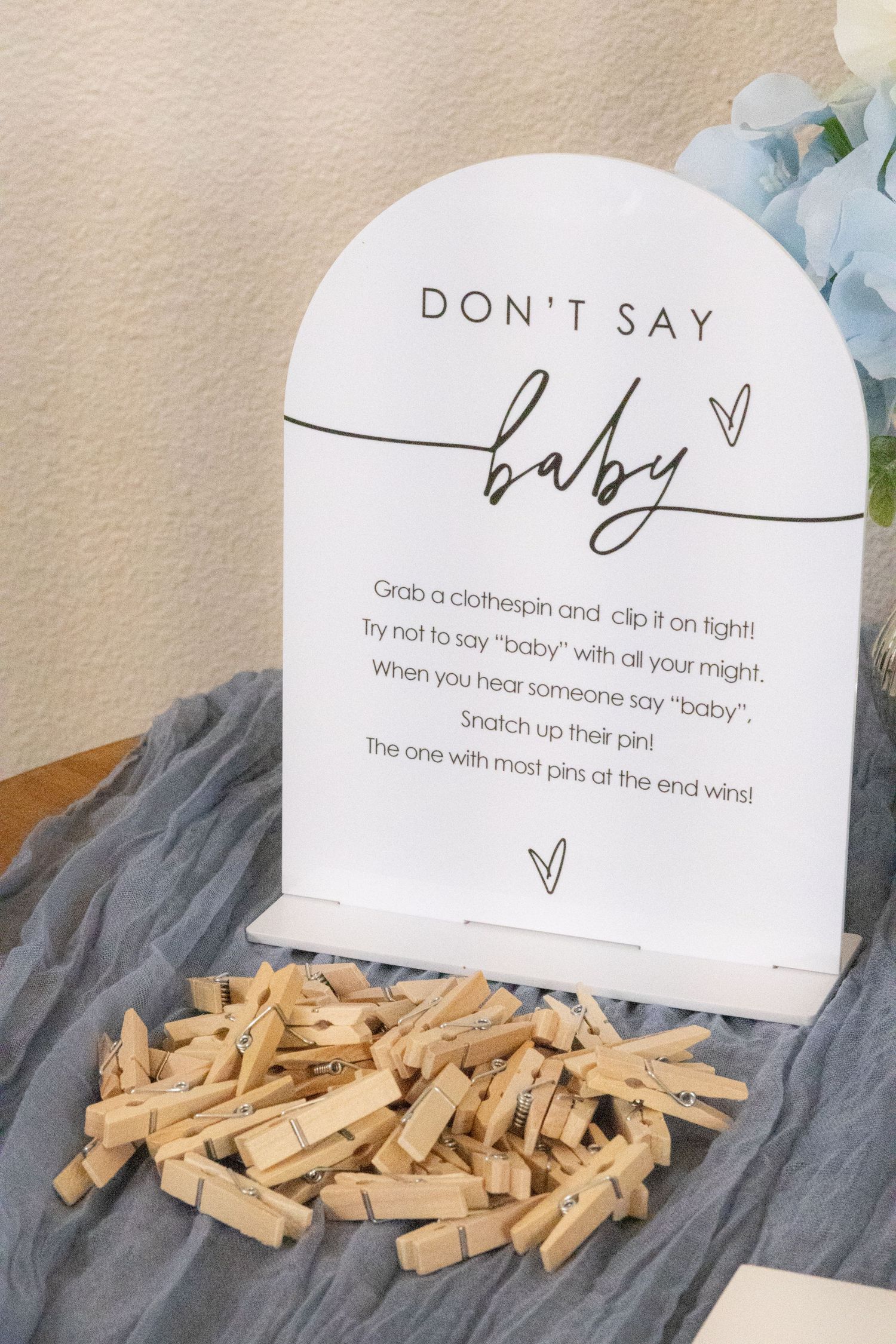 Don't Say Baby shower game sign with a pile of wooden clothespins on a blue cloth beside blue flowers.