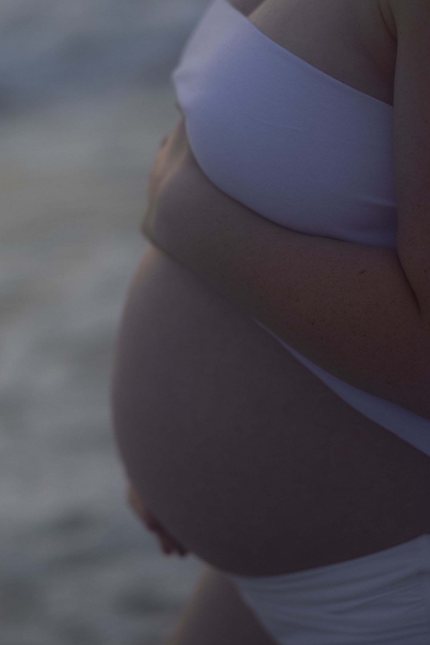 Soft, moody close-up of a pregnant belly in a white two-piece outfit beside the ocean at dusk.