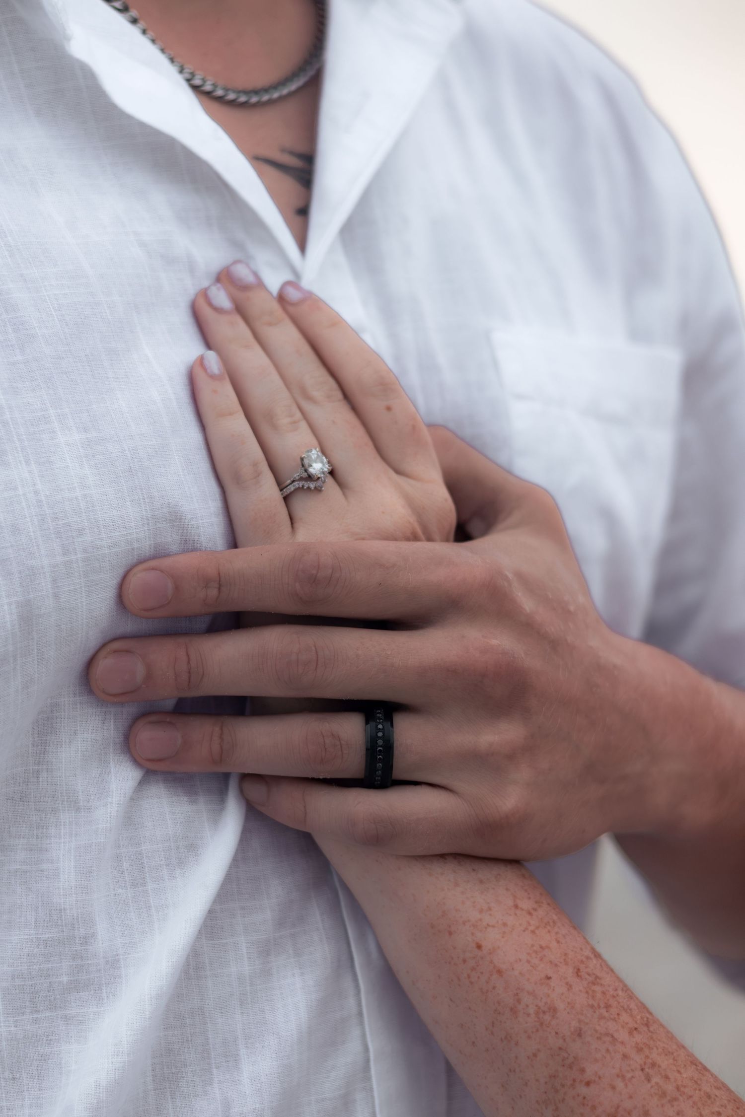Close-up of a woman's hand with a diamond engagement ring resting on a man's white linen shirt, his black wedding band visible.