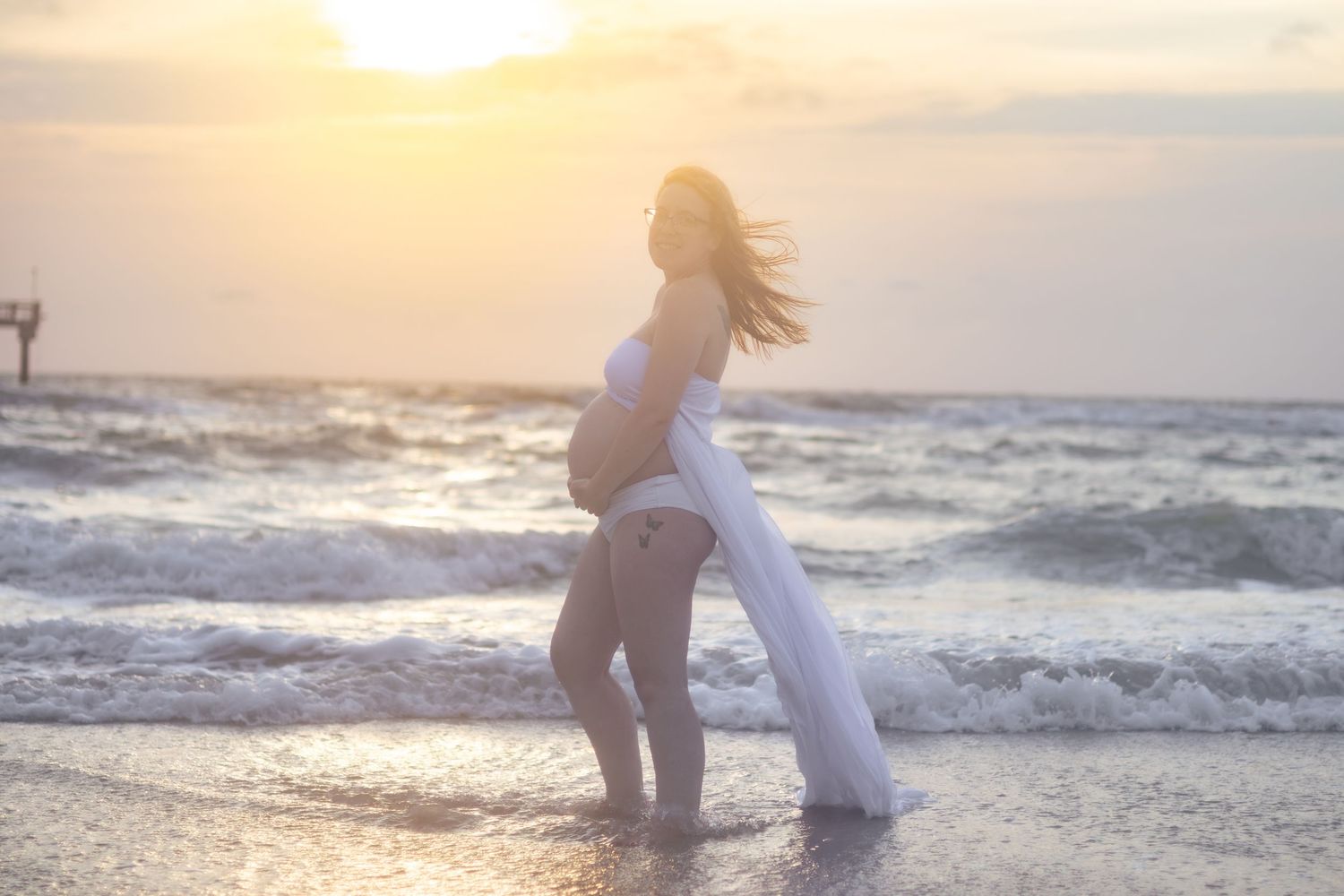 Pregnant woman in a white gown stands in shallow ocean waves with her hair blowing in the breeze at a golden sunset.