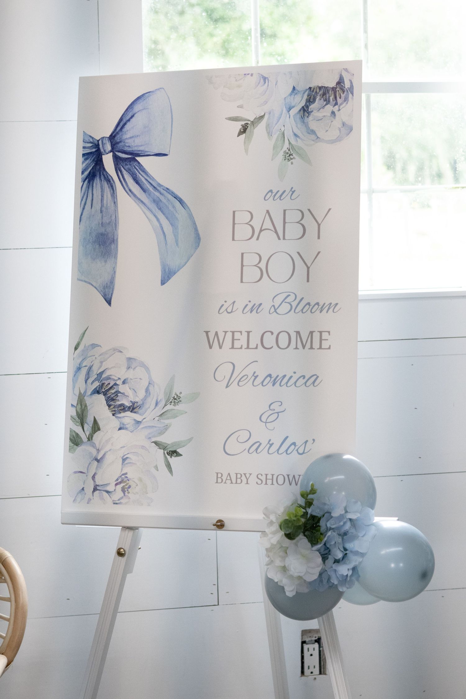 Baby shower welcome sign with blue bow and floral design reading baby boy is in bloom, beside blue balloons.