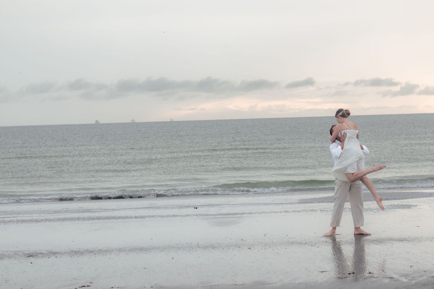 Couple shares a romantic kiss and lift on a wide empty beach with calm ocean waters and a soft cloudy sky behind them.