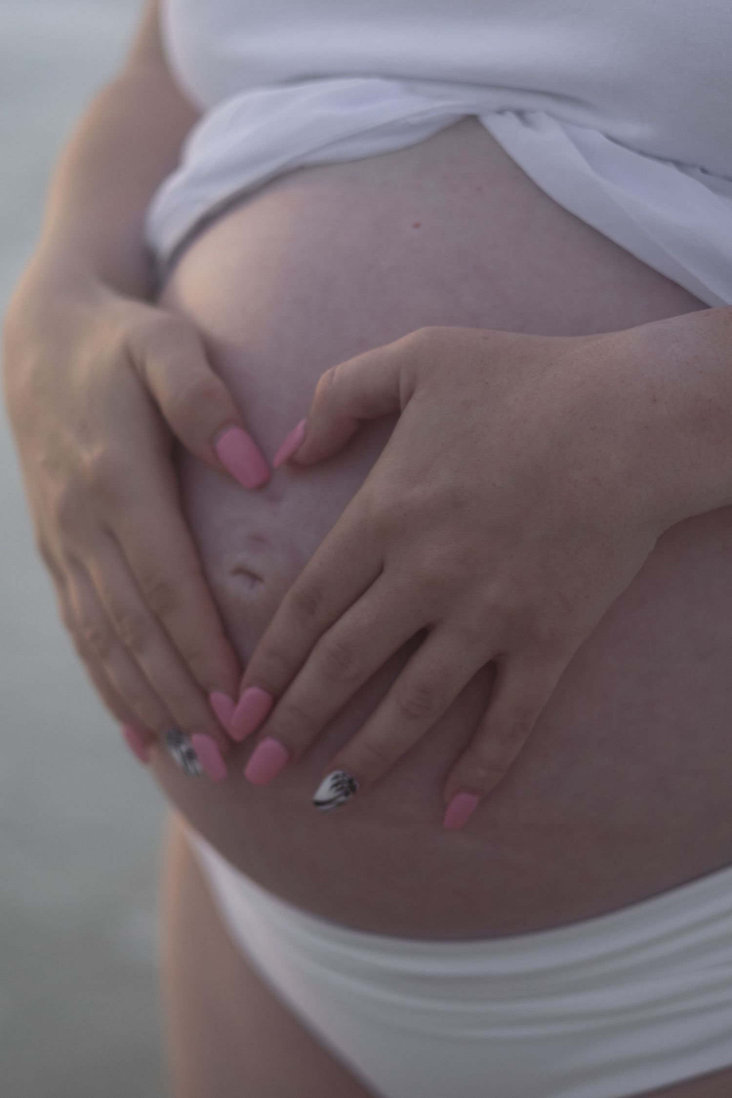 A pregnant woman with pink nails forms a heart shape with her hands on her bare baby bump at the beach.