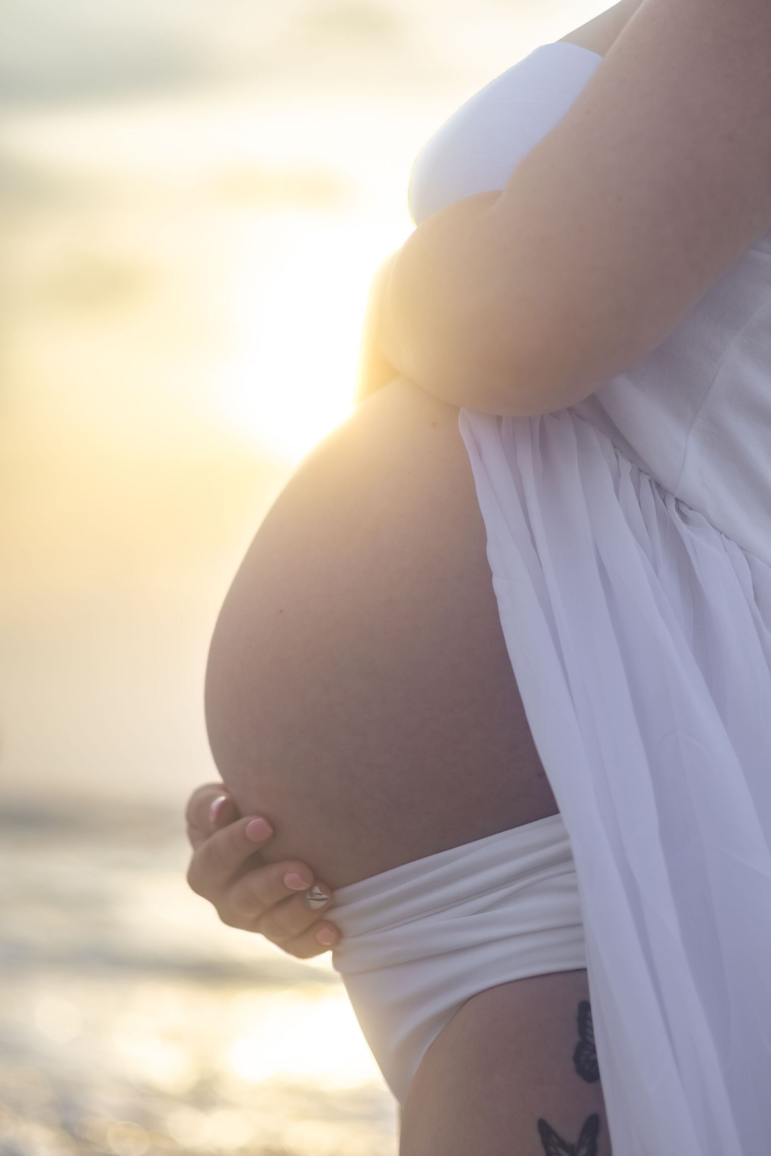 Close-up of a pregnant belly glowing in golden sunset light as a woman in white cradles her bump on the beach.