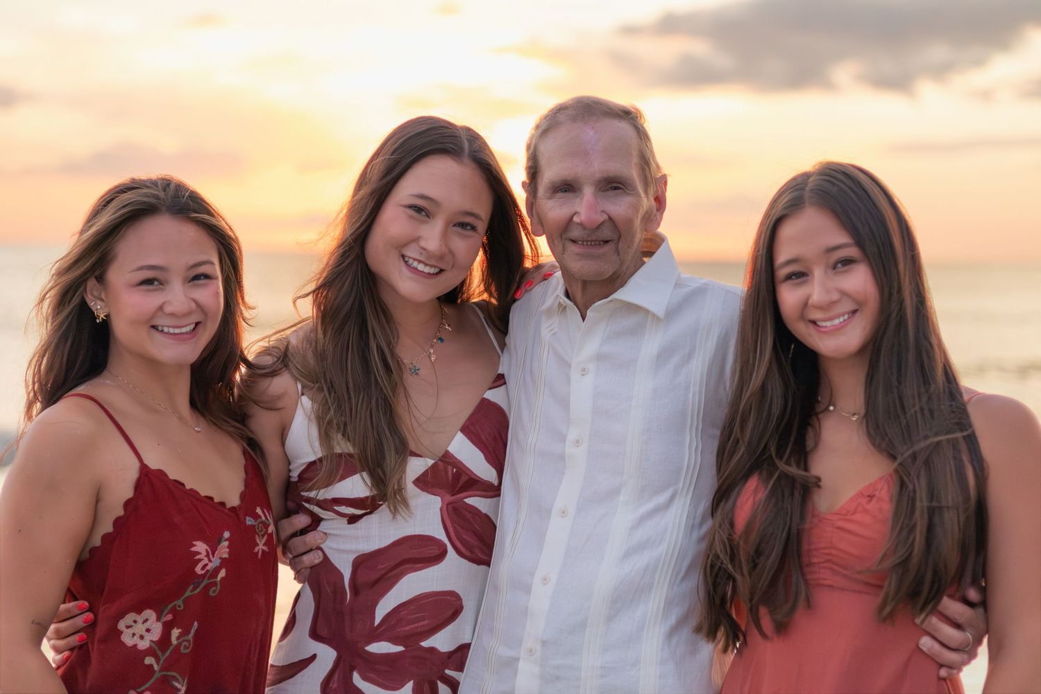 Close-up sunset beach portrait of a smiling older man standing arm in arm with three women in red and floral dresses.
