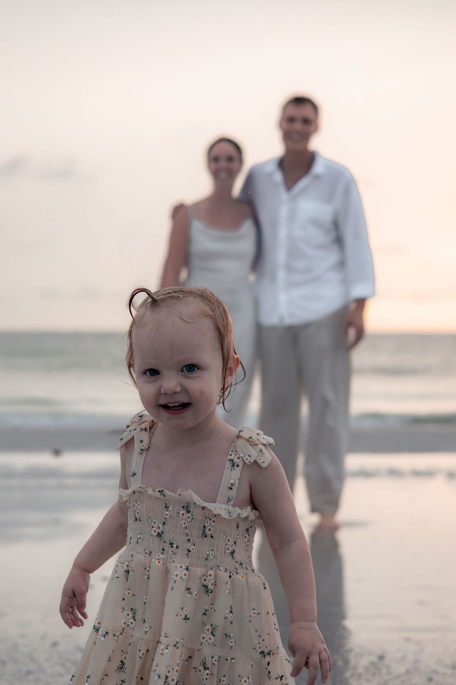 Smiling toddler in a floral dress runs along a beach at sunset while two blurred parents stand in the shallow water behind her.
