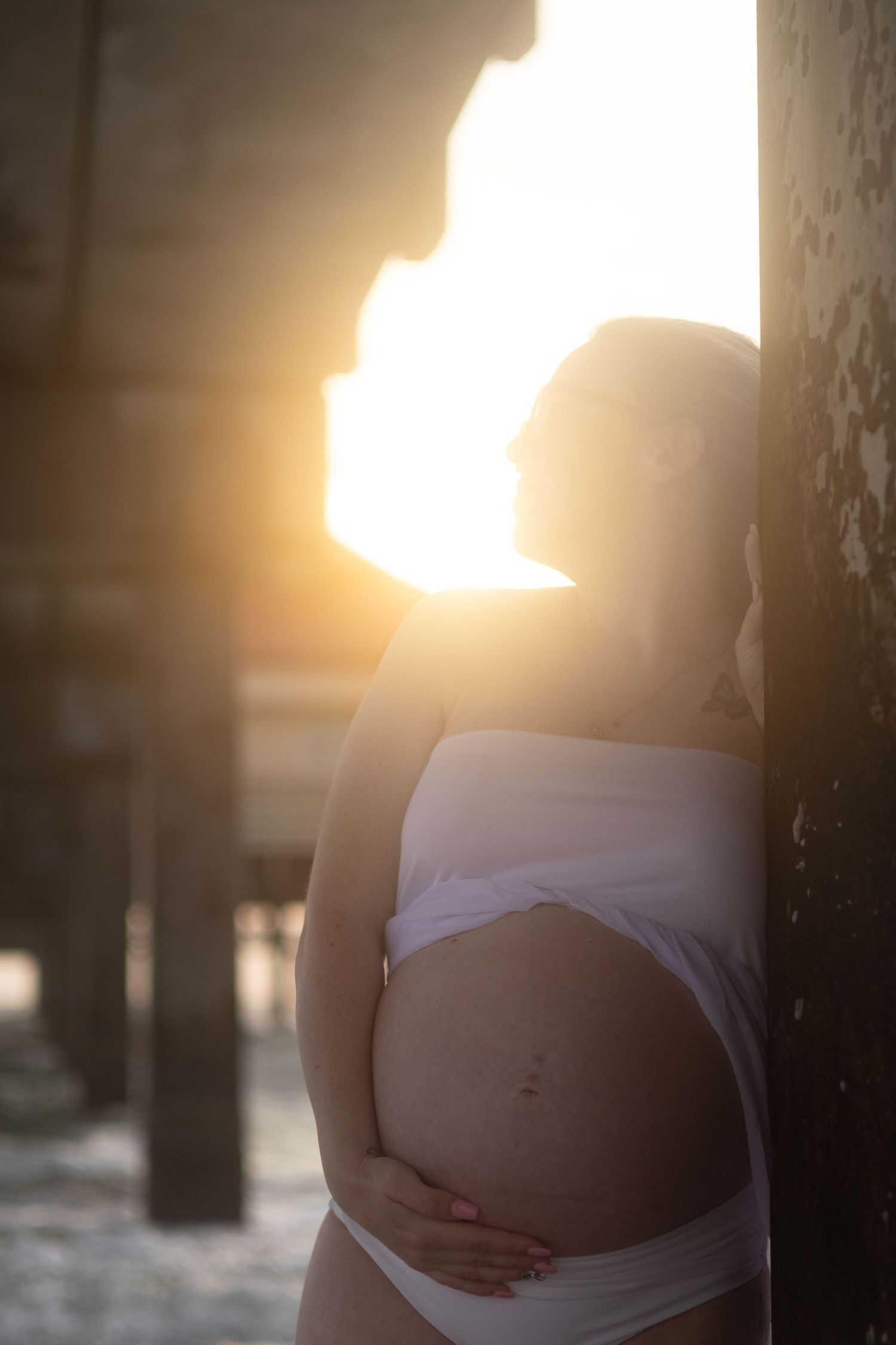 Pregnant woman in a white wrap leans against a weathered pier post as golden sunset light glows behind her.