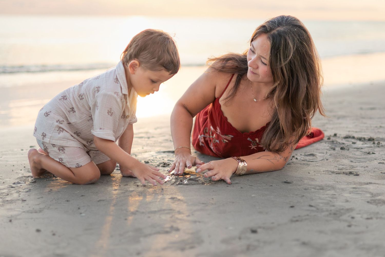 A smiling mother and her young son play together in the wet sand at the beach while the sun sets behind them.
