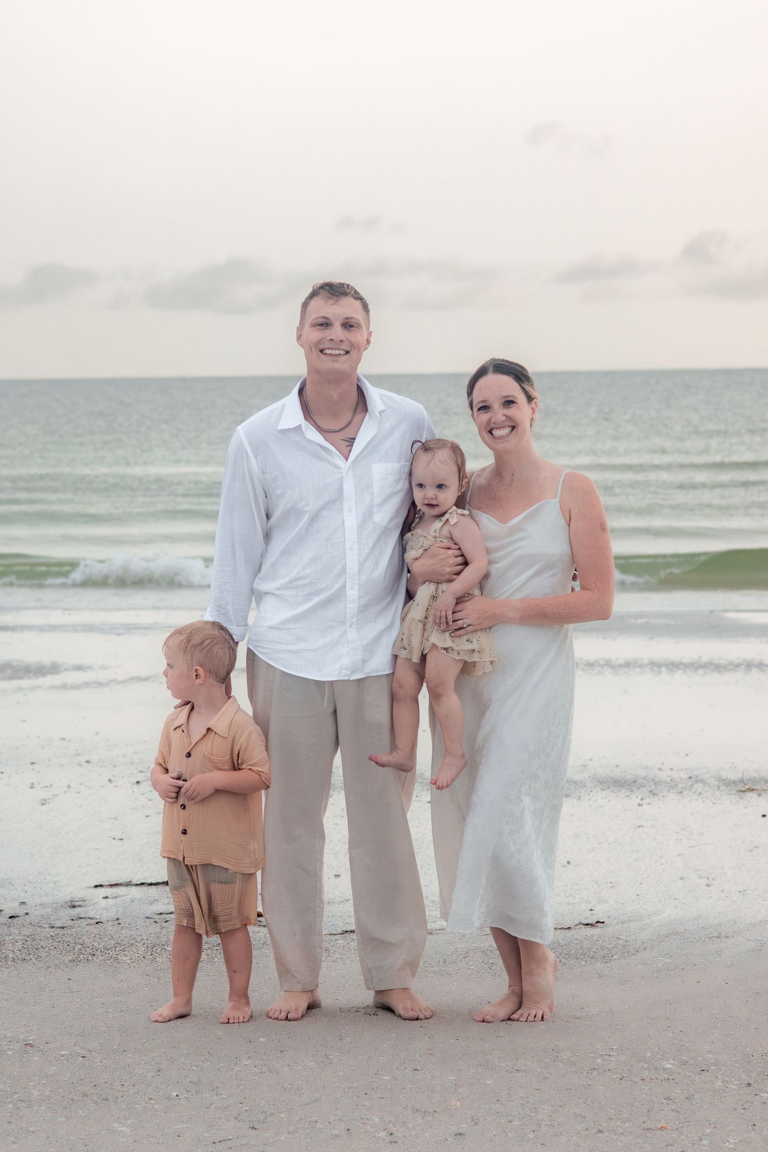 Family of four in white and neutral linen outfits standing barefoot on a sandy beach with ocean waves and a soft sunset sky behind them.