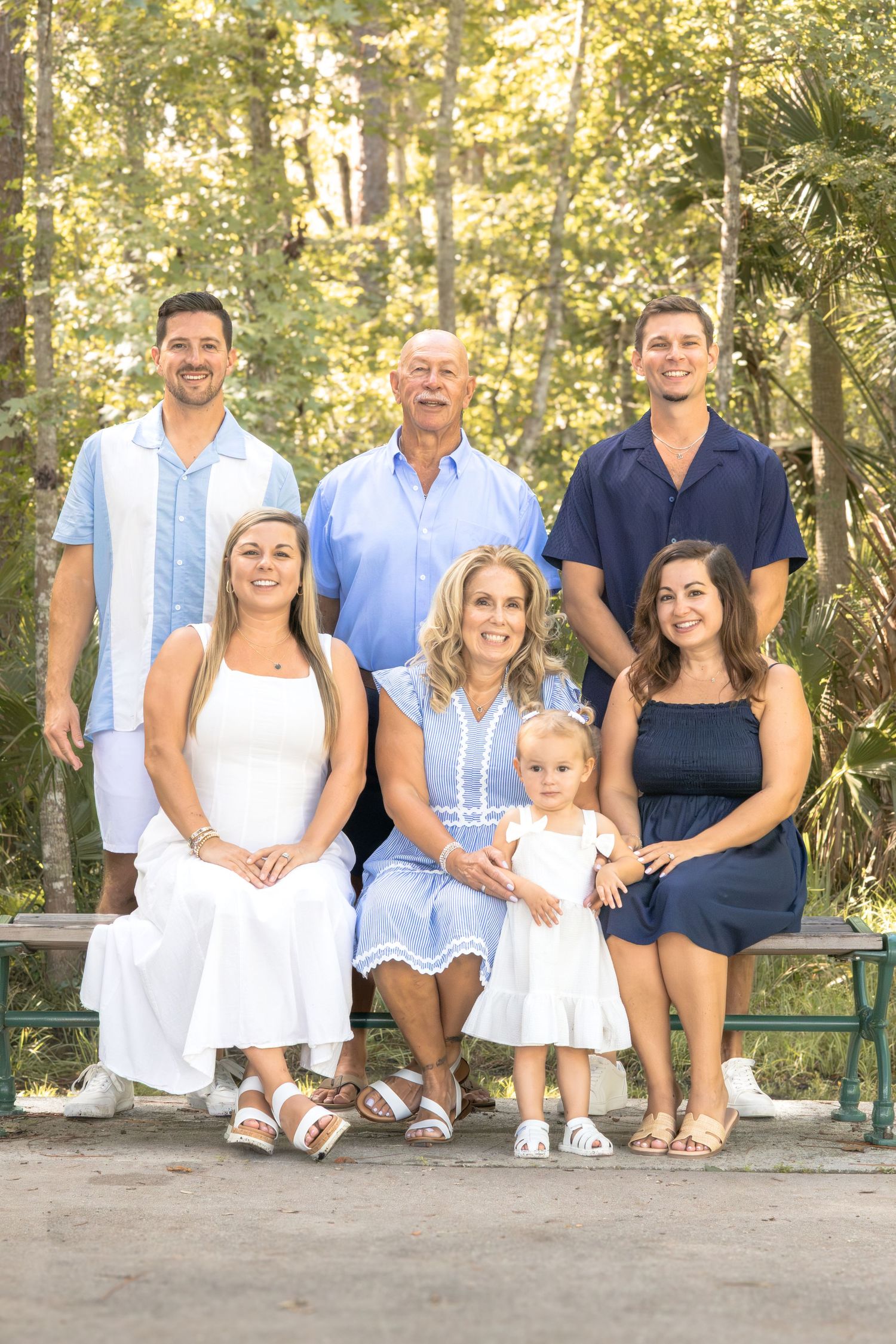 Family of six in coordinated blue and white outfits posing outdoors, three men standing behind two women and a toddler seated on a bench.