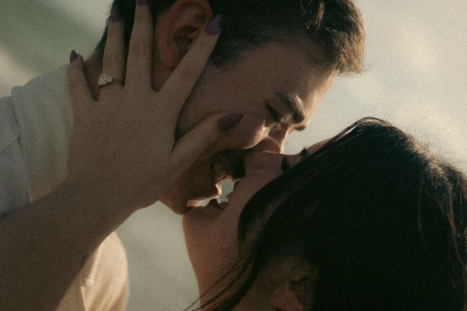 Close-up of a romantic couple sharing an intimate kiss, her hand with a ring and purple nails gently touching his face in soft light.