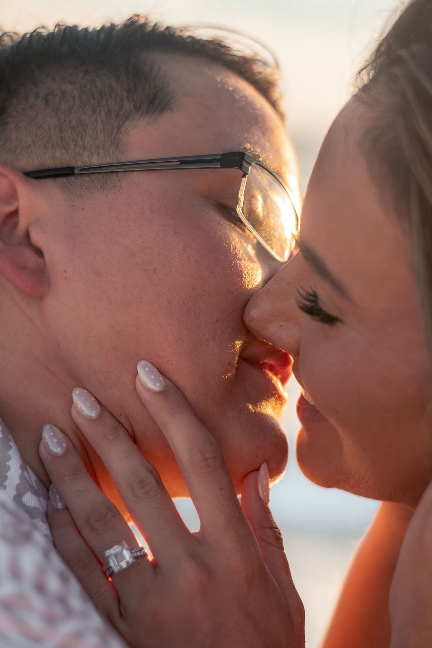Close-up of two people about to kiss at sunset, one wearing glasses, with a hand showing a diamond engagement ring and light manicure.