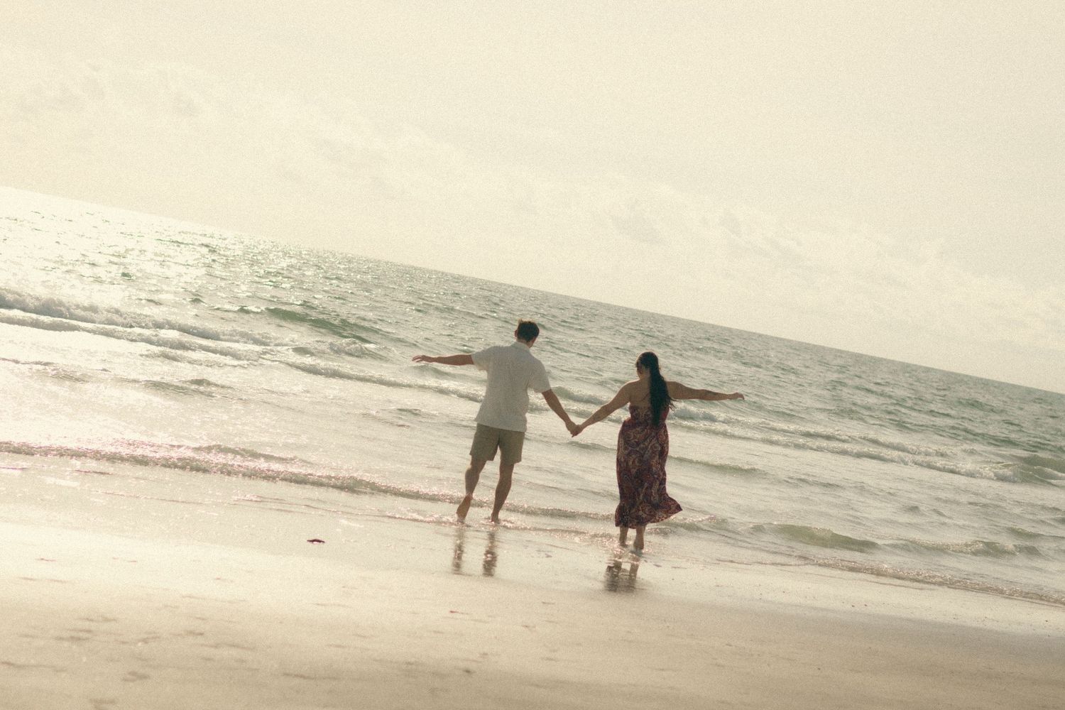 Couple holding hands walk barefoot along the wet sand of a calm beach at sunset, with gentle waves rolling in under a soft hazy sky.
