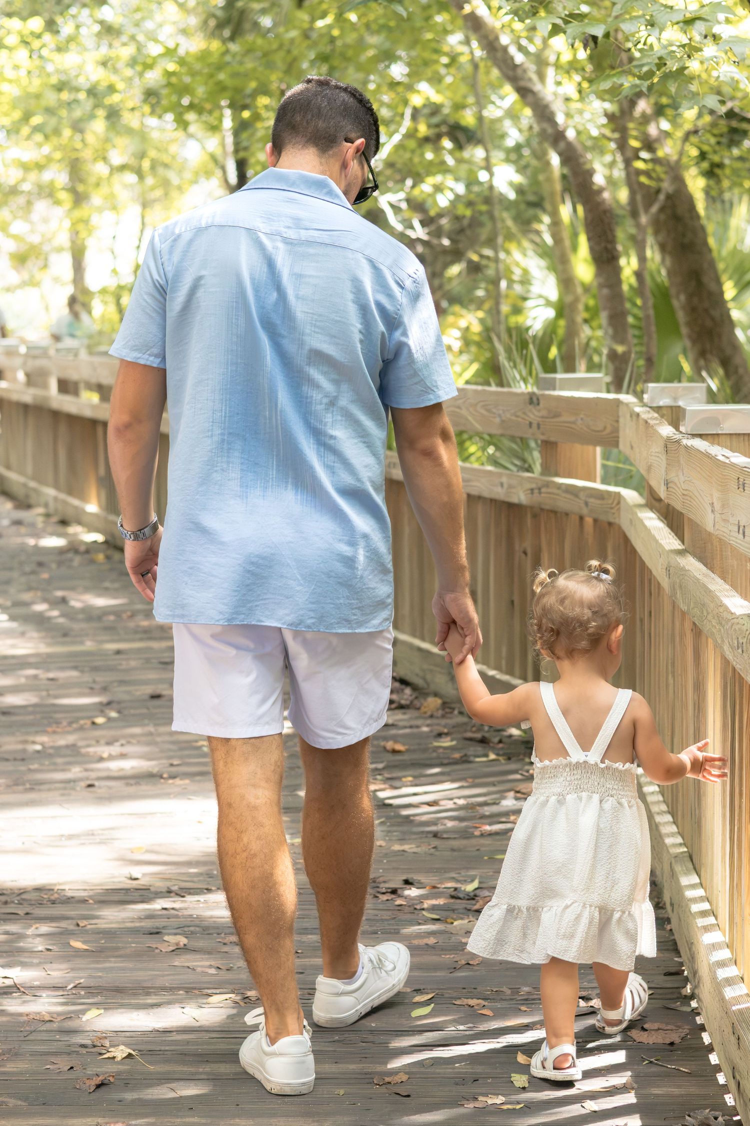 Father in light blue shirt and white shorts holds hands with a toddler in a white dress while walking along a wooden boardwalk.