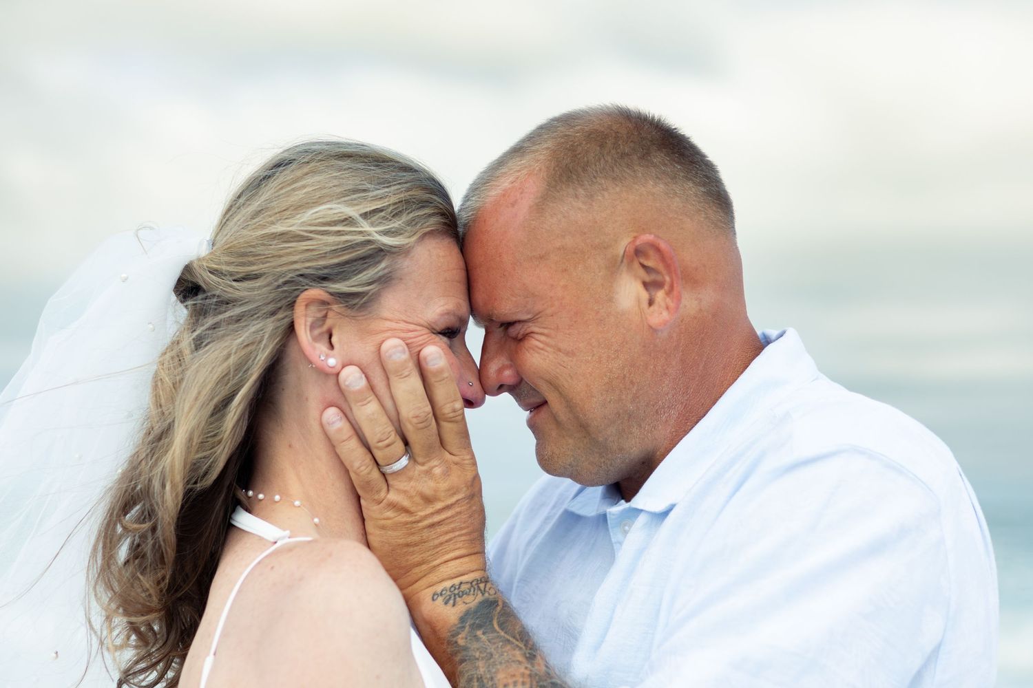 Bride in a white veil and groom in a light blue shirt touch foreheads while she cradles his face on a beach wedding day.