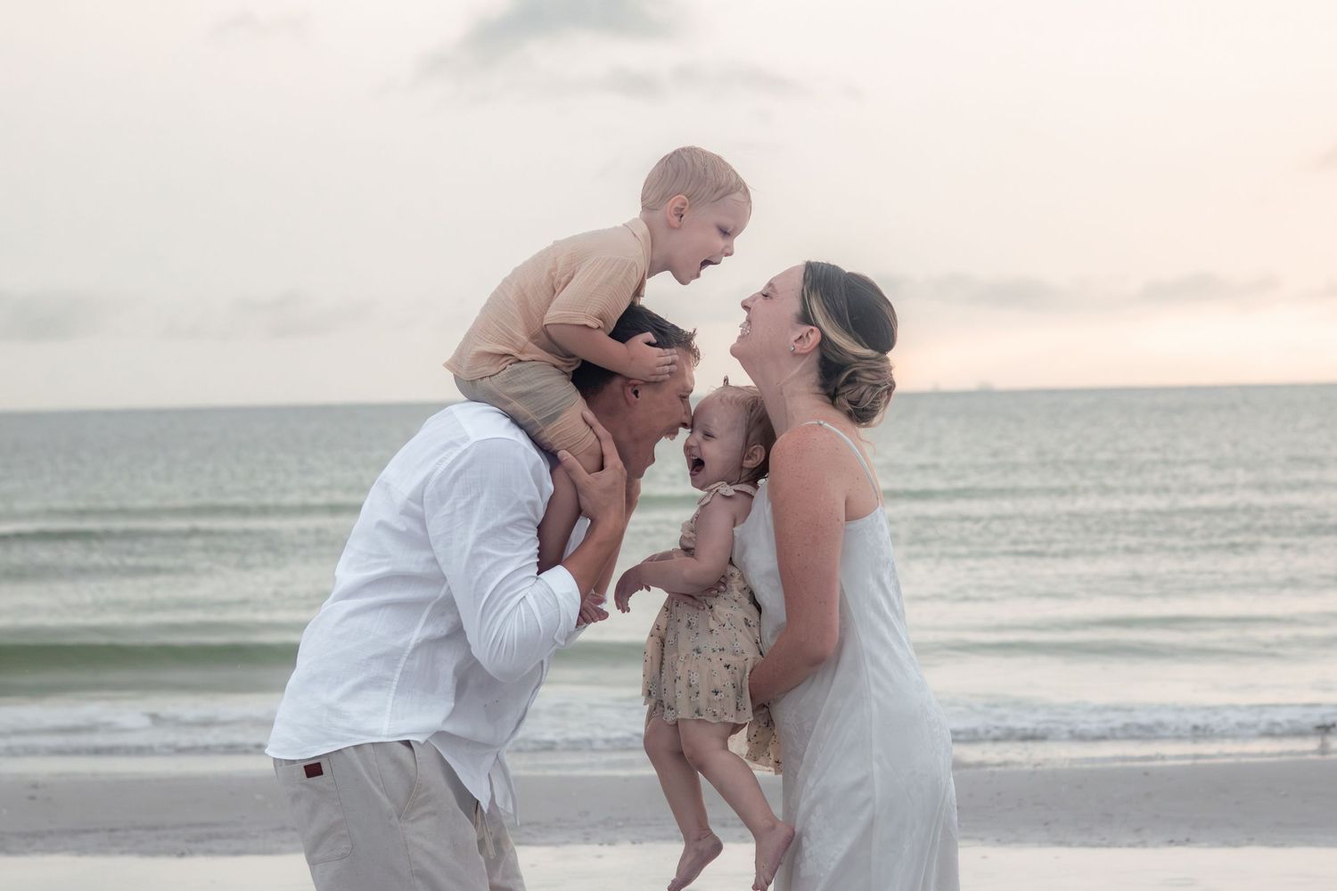 A family of four shares a joyful moment on the beach at sunset, with a father holding a toddler on his shoulders.