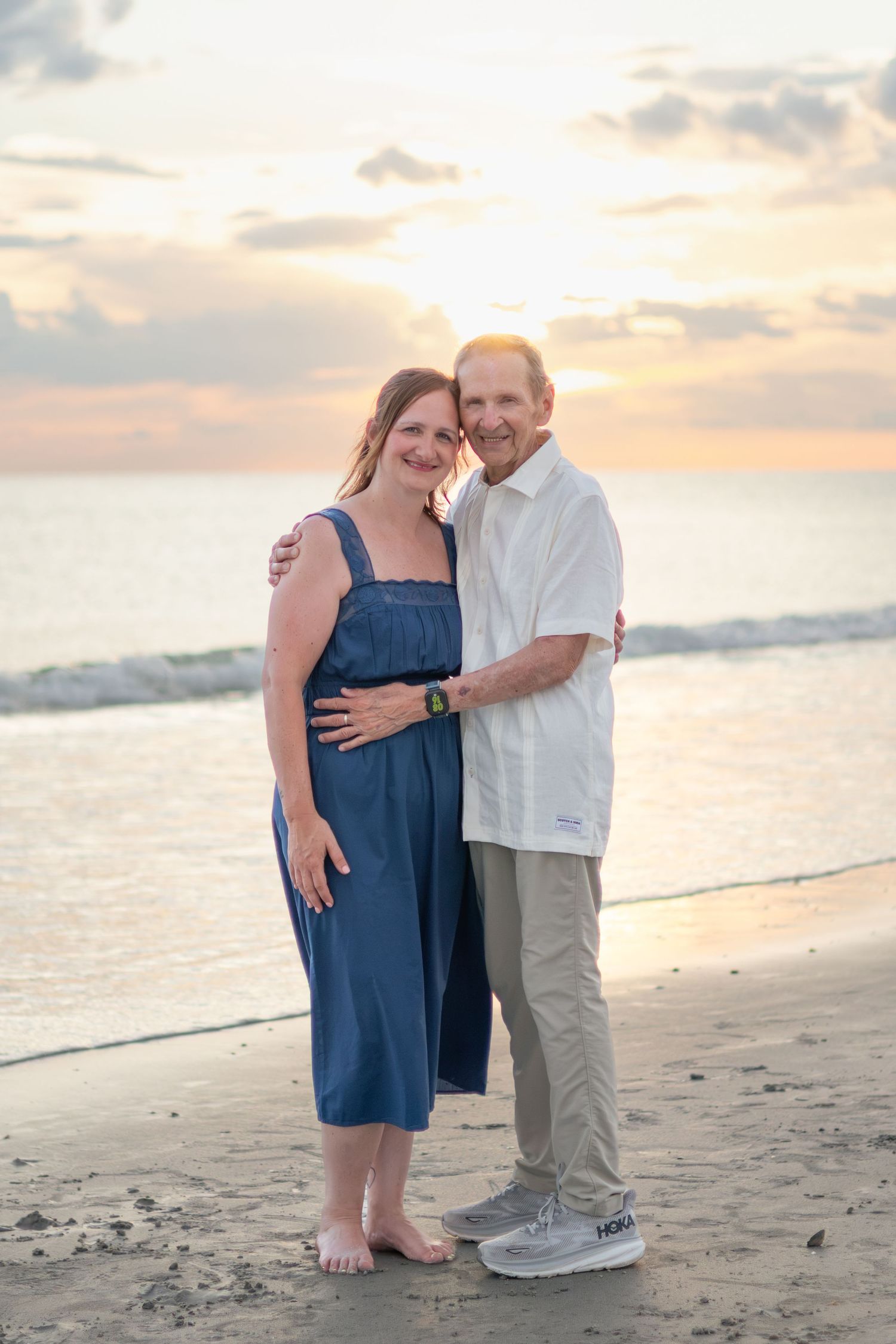 A happy couple embraces on the beach at sunset, smiling at the camera as gentle waves roll in behind them.