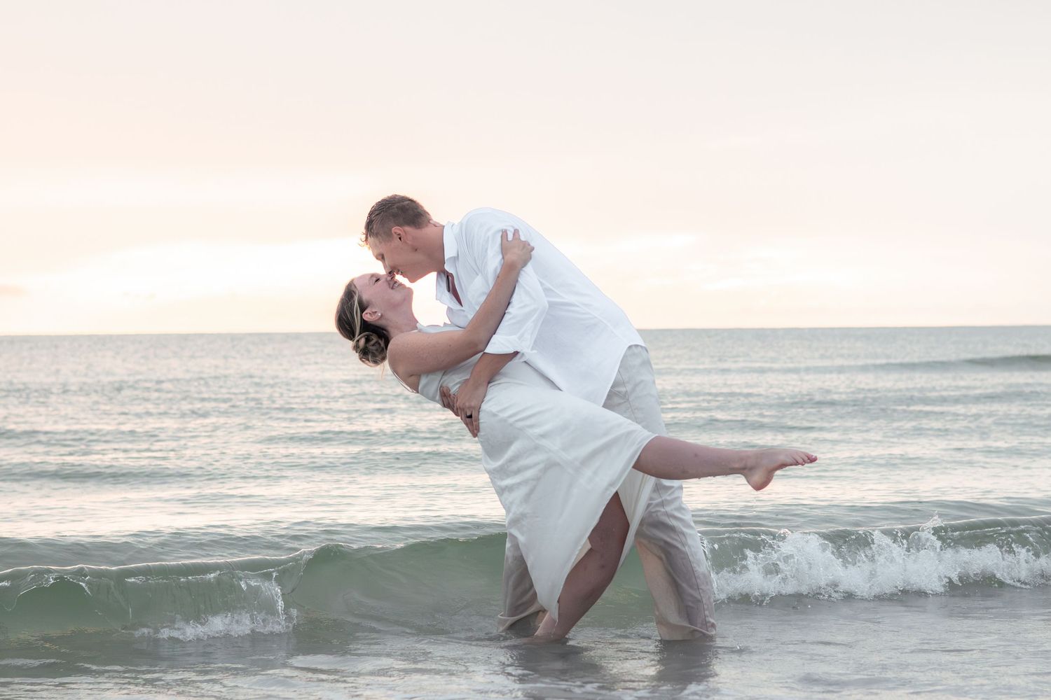 Bride in a white dress is dipped backward for a romantic kiss in the ocean waves at sunset.