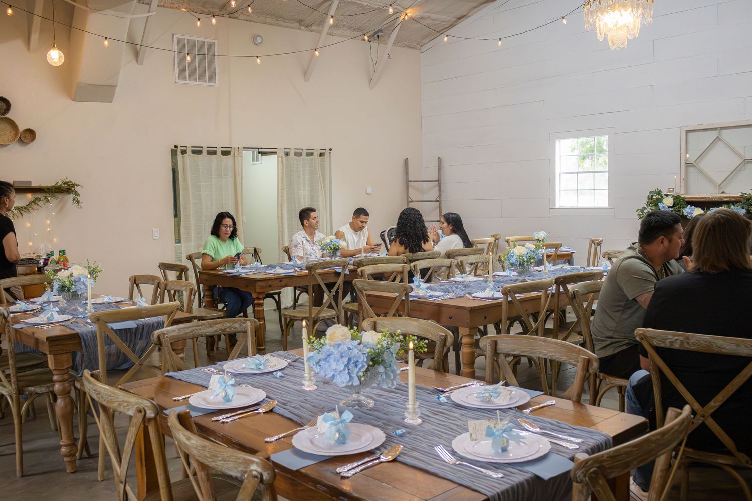 Guests seated at rustic wooden tables in a bright venue, set with blue floral centerpieces and candles for a baby shower.