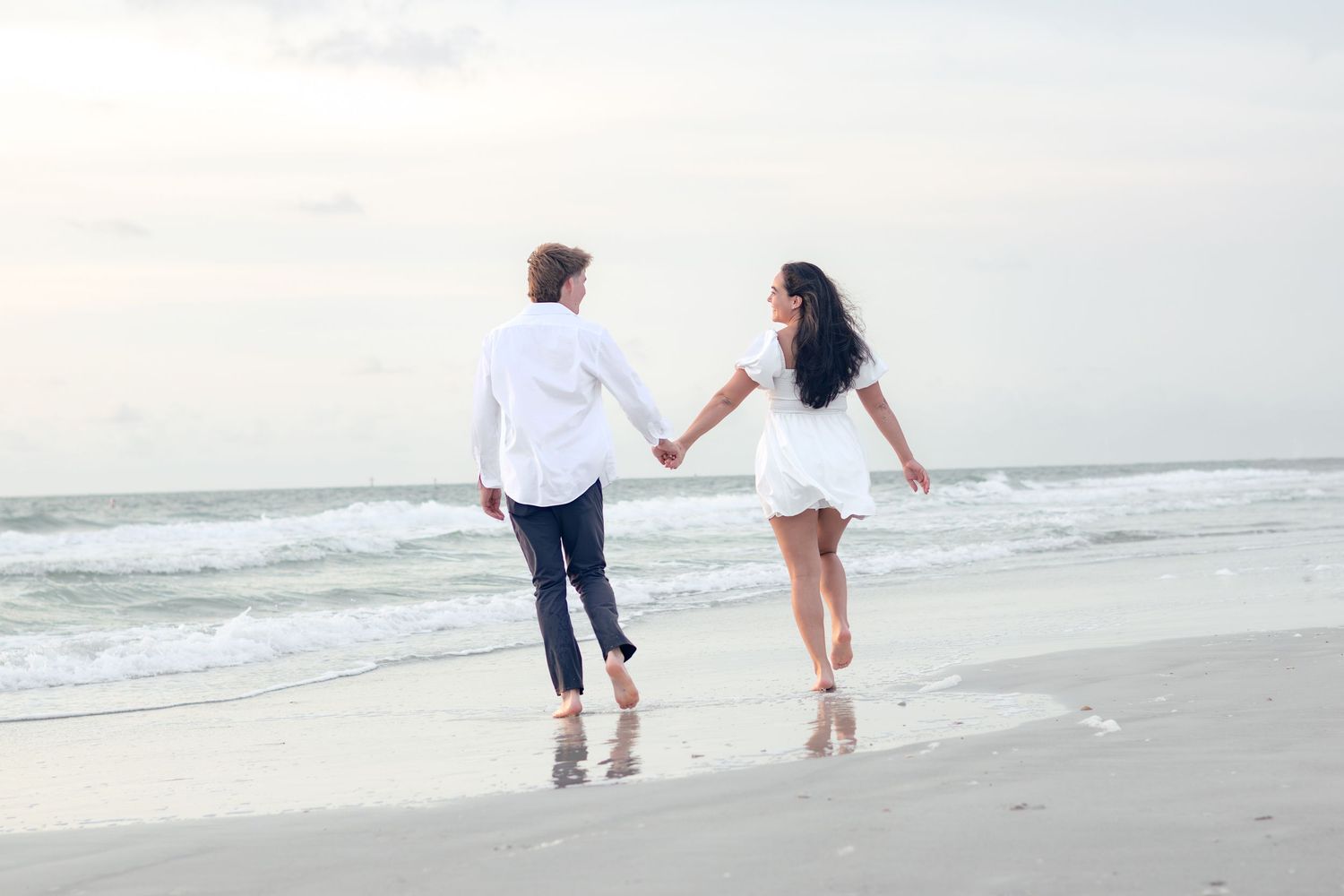 Barefoot couple holding hands while strolling along the wet sand of a beach as waves roll in.