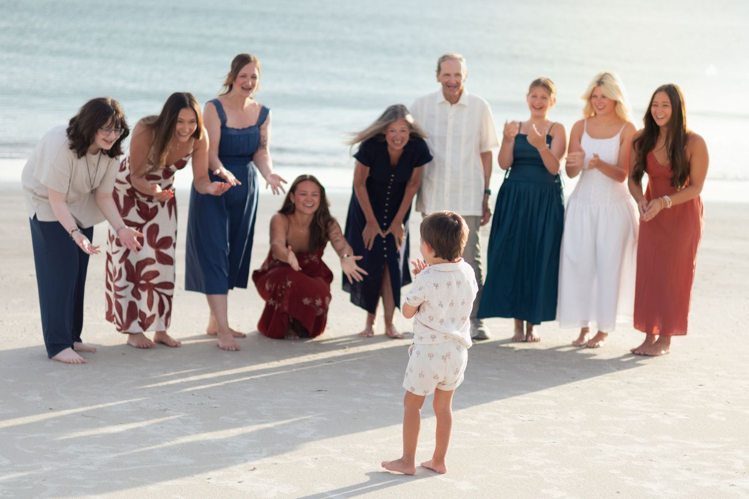 Group of women and a man smile and gather on the beach as a toddler in white stands in front during a family photo session.