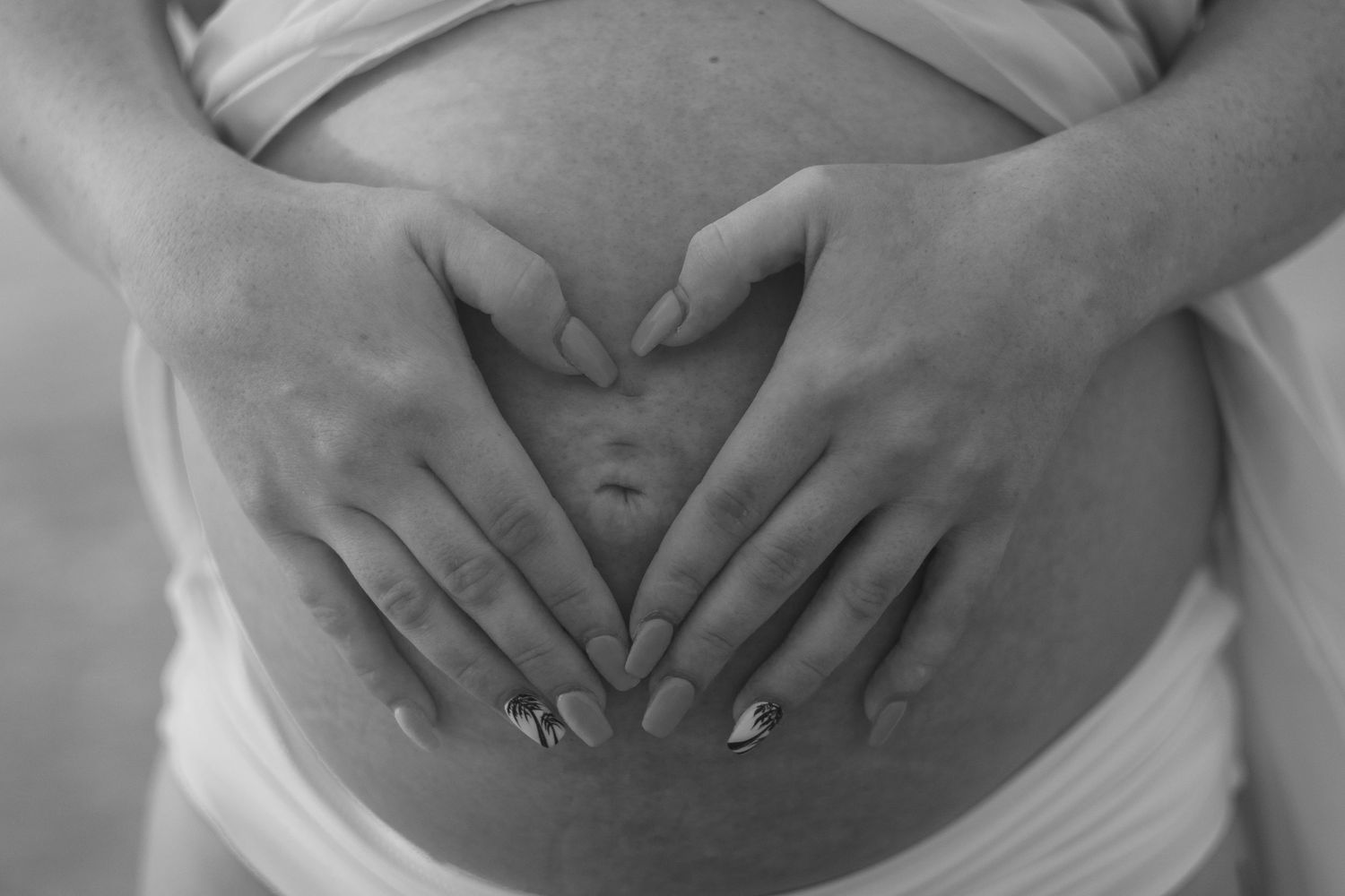 Black and white close-up of a pregnant woman's hands forming a heart shape on her bare baby bump.