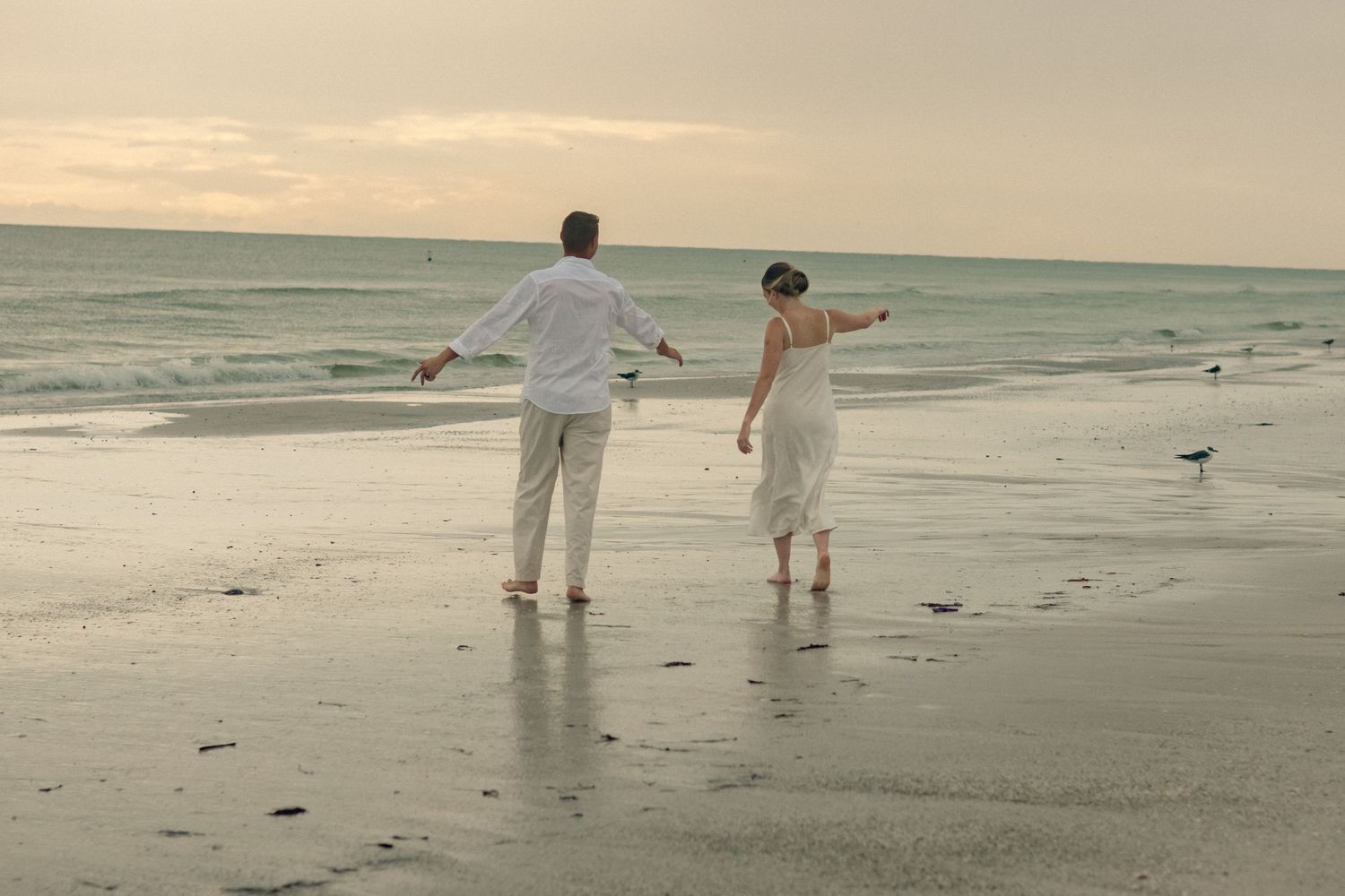 A couple in white clothing stands on the wet sand with arms spread wide, facing the ocean as shorebirds linger nearby.