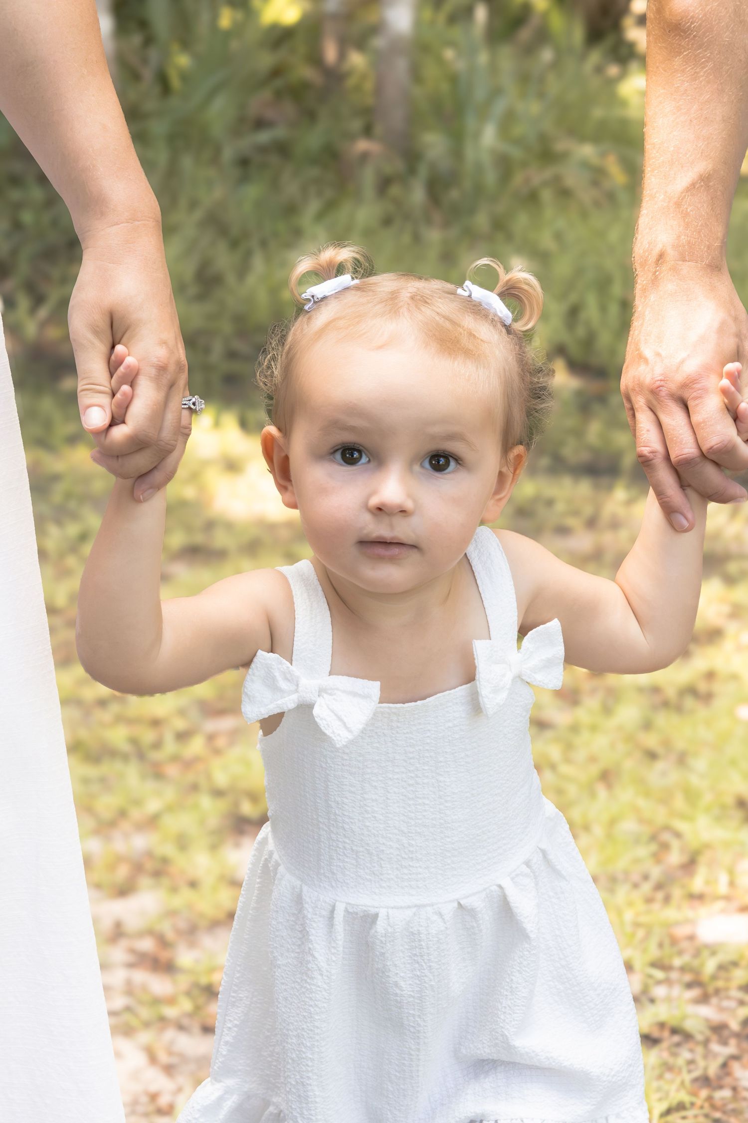 Toddler girl in a white bow dress and hair clips holds two adults' hands while walking outdoors through a sunlit park.