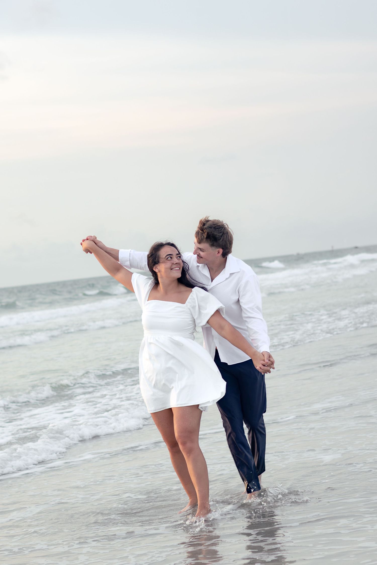 Smiling couple holding hands and playfully twirling in shallow ocean water in white dress and shirt.
