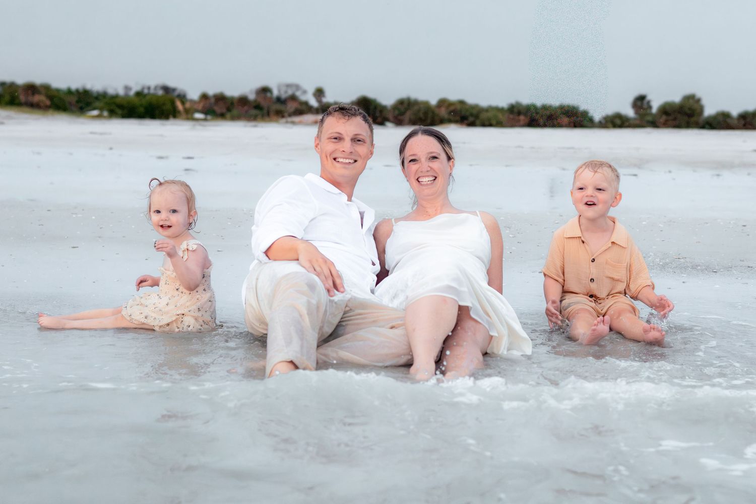 Family of four in neutral outfits sits together in shallow ocean water at the beach, smiling for a coastal family portrait.