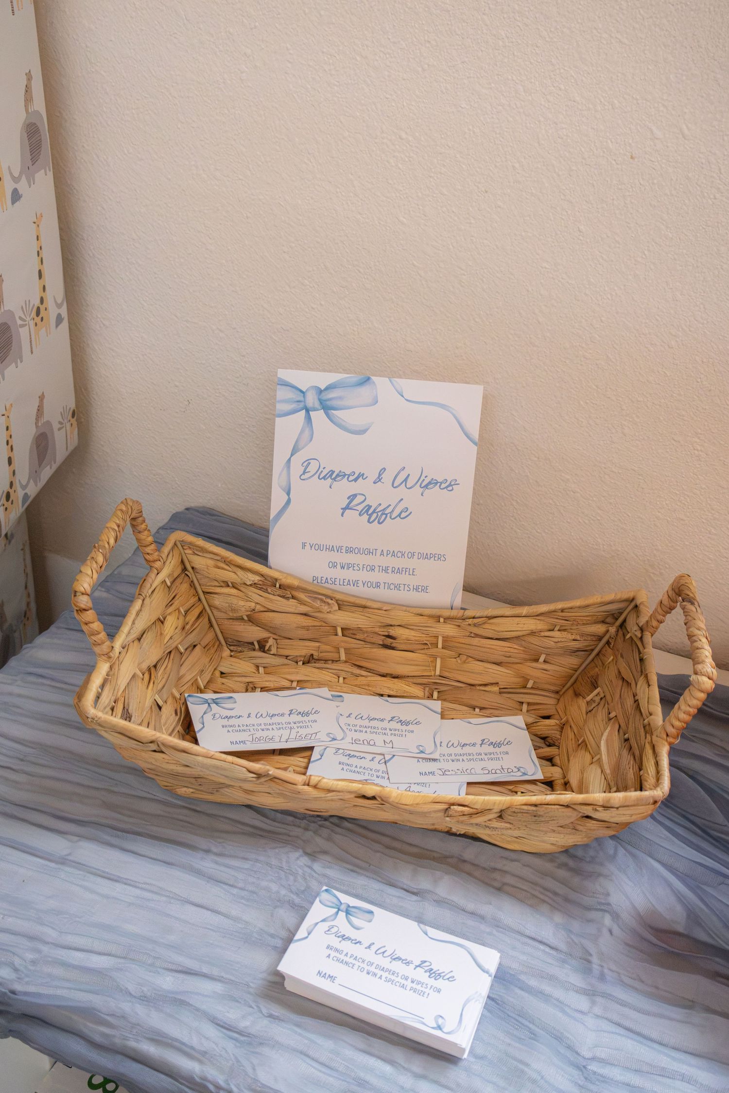 Wicker basket with diaper and wipes raffle tickets beside a blue bow sign on a gray runner at a baby shower.