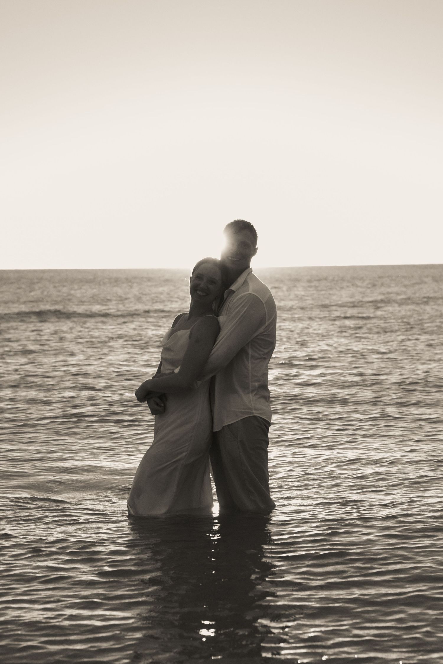 Sepia silhouette of a couple embracing in the sea as the sun glows behind them on the horizon.