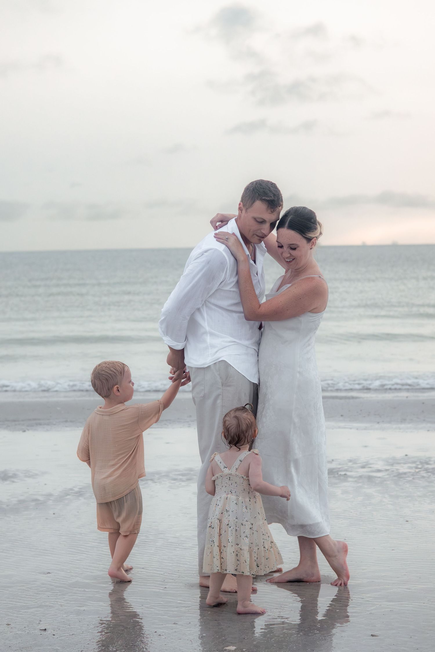Parents embrace on the beach in white and neutral clothing while a young boy and toddler girl hold their hands by the ocean.